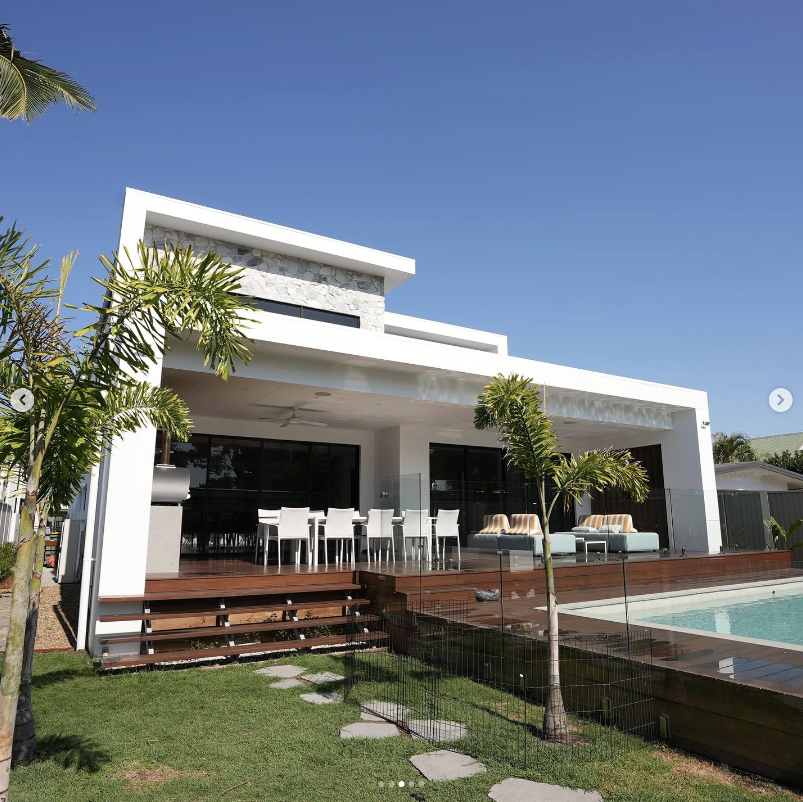 Modern white house with a flat roof, large windows, an outdoor dining area with white chairs, a sitting area with striped cushions, a wooden deck, a swimming pool, and lush green trees, under a clear blue sky.