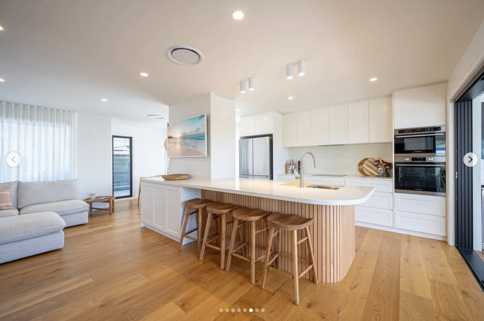Modern open-concept kitchen with white cabinetry, marble countertops, and a wooden island with four bar stools, adjacent to a living area with a white sofa and a large window with sheer curtains.