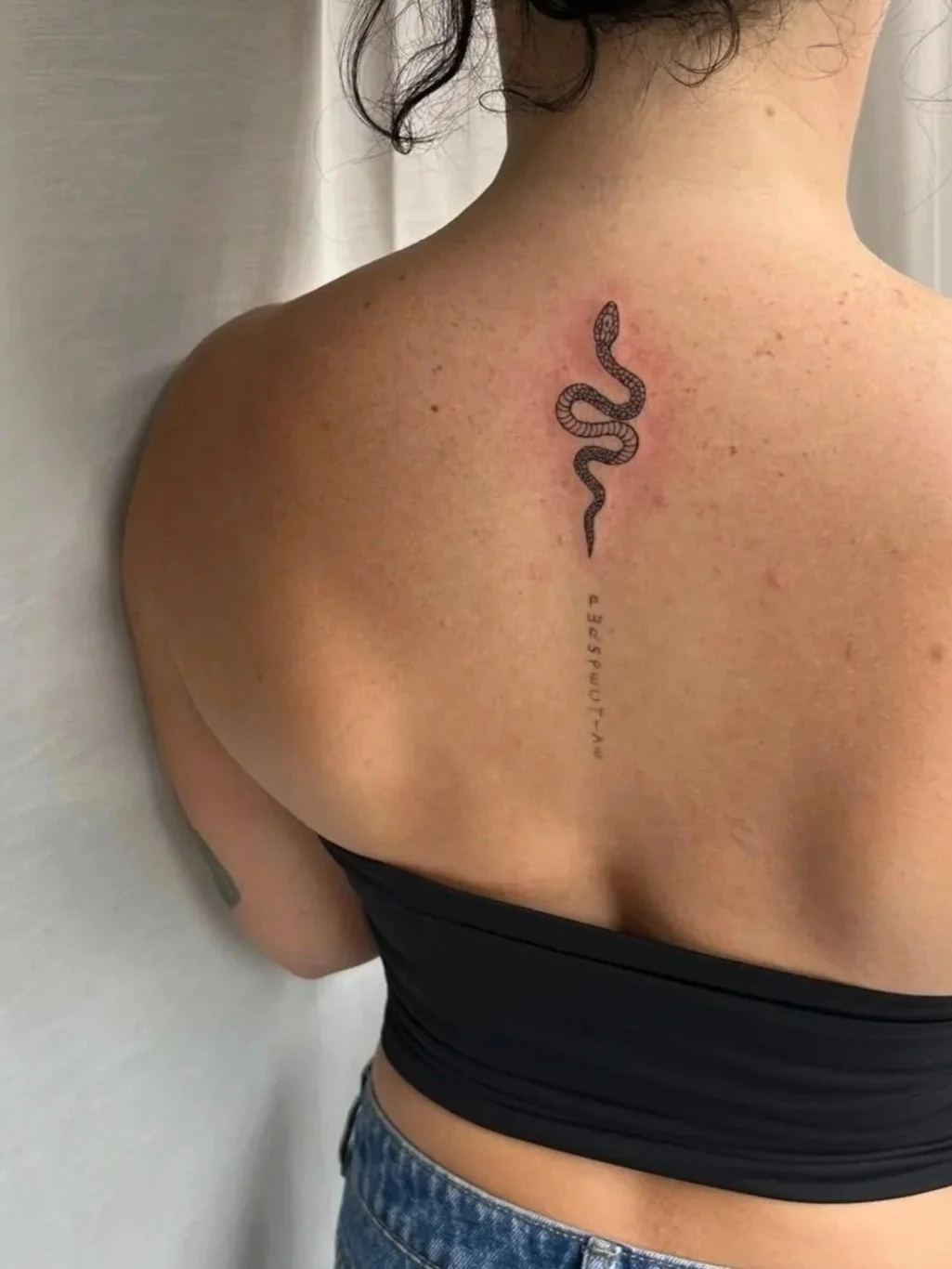 Back of a person with a fine line snake tattoo running down the upper spine.
