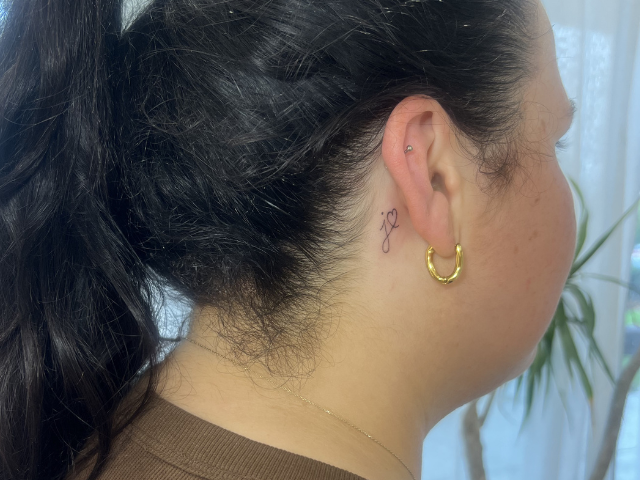 Ladies side profile with fine line tattoo behind ear of a J with heart