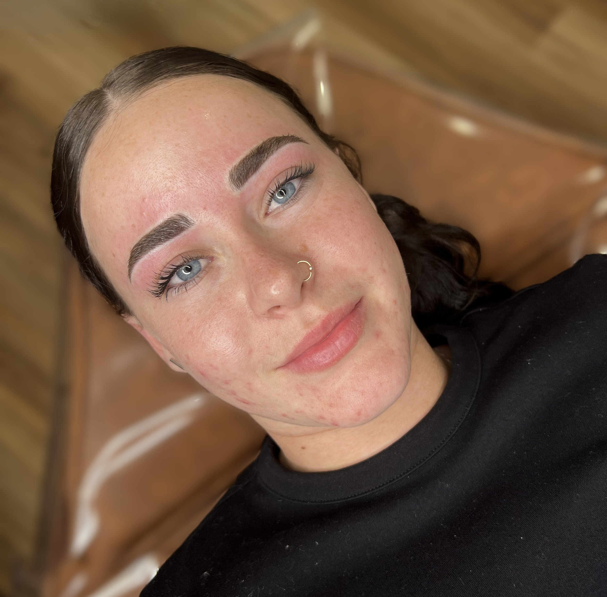 Close-up photo of a woman's face with recently completed eyebrow microblading, light skin with freckles, blue eyes, and dark hair, lying on salon chair.