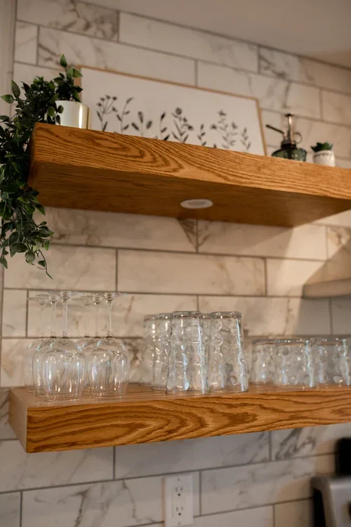Open wooden kitchen shelves at the Villa at Ramble Creek, displaying glassware, small plants, and decor against a marble-style tile backsplash.