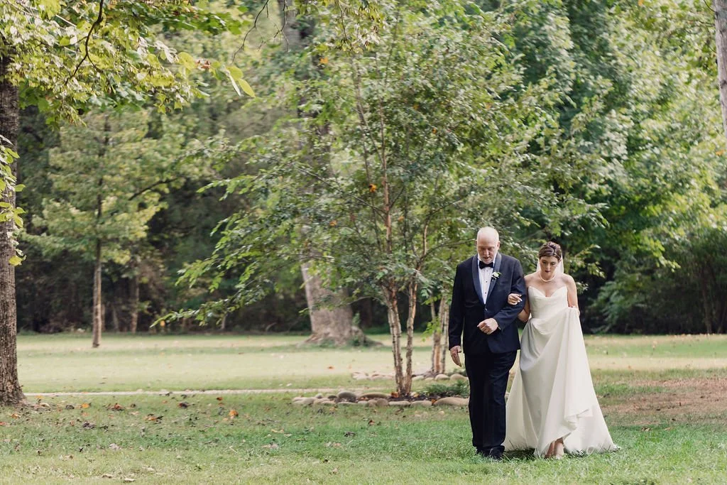 A bride walking down the aisle with her father at Ramble Creek wedding venue with green trees and grass.