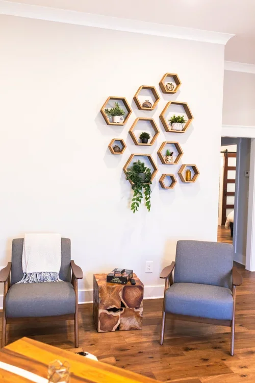 Living room with two grey armchairs, a wooden side table, and a wall with hexagonal wooden shelves holding small plants and decorative objects. One of three accommodations at Ramble Creek Tennessee wedding venue with on-site lodging.