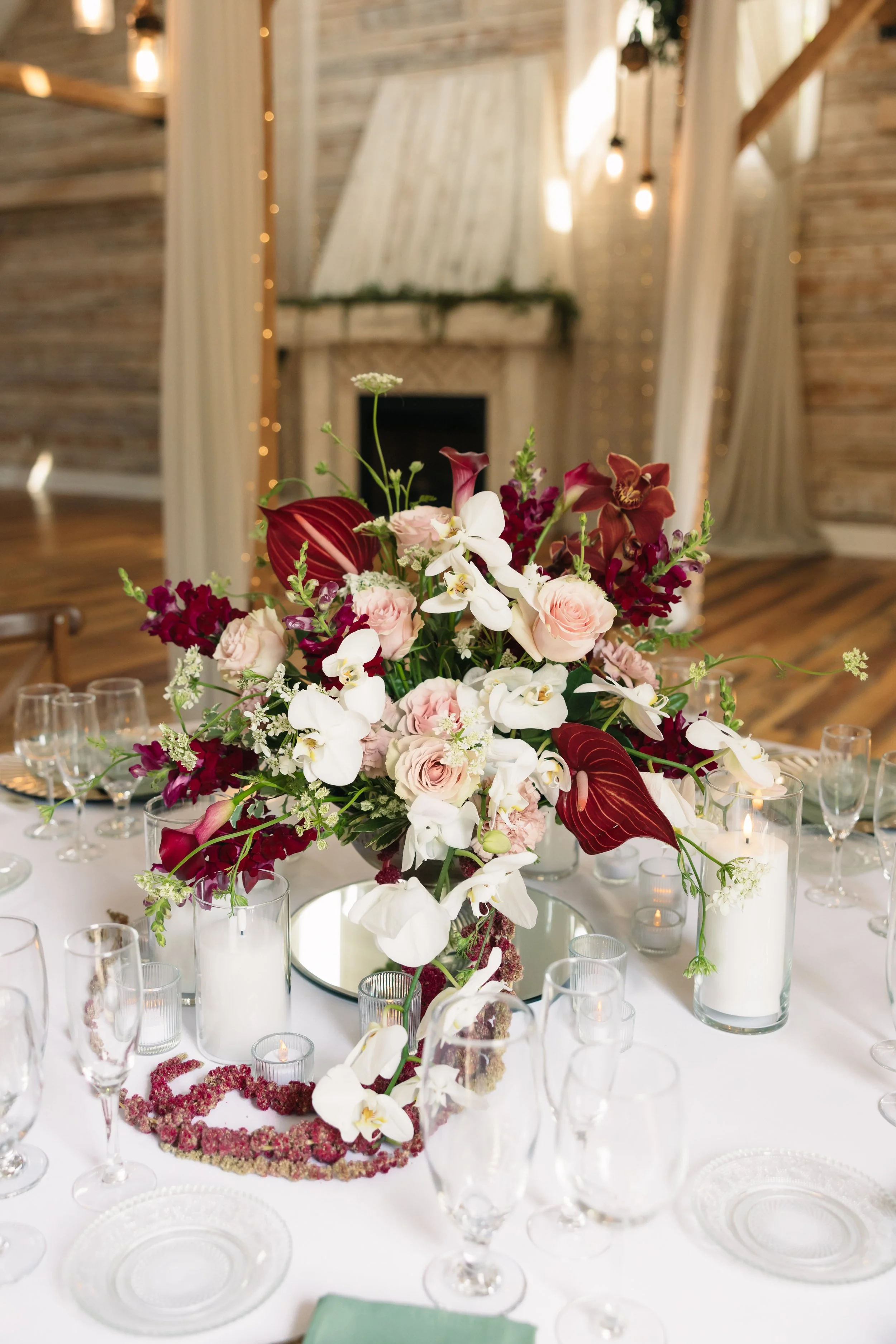 Wedding reception centerpiece at Ramble Creek featuring burgundy orchids, blush roses, white florals, candlelight, glassware, and elegant draping.