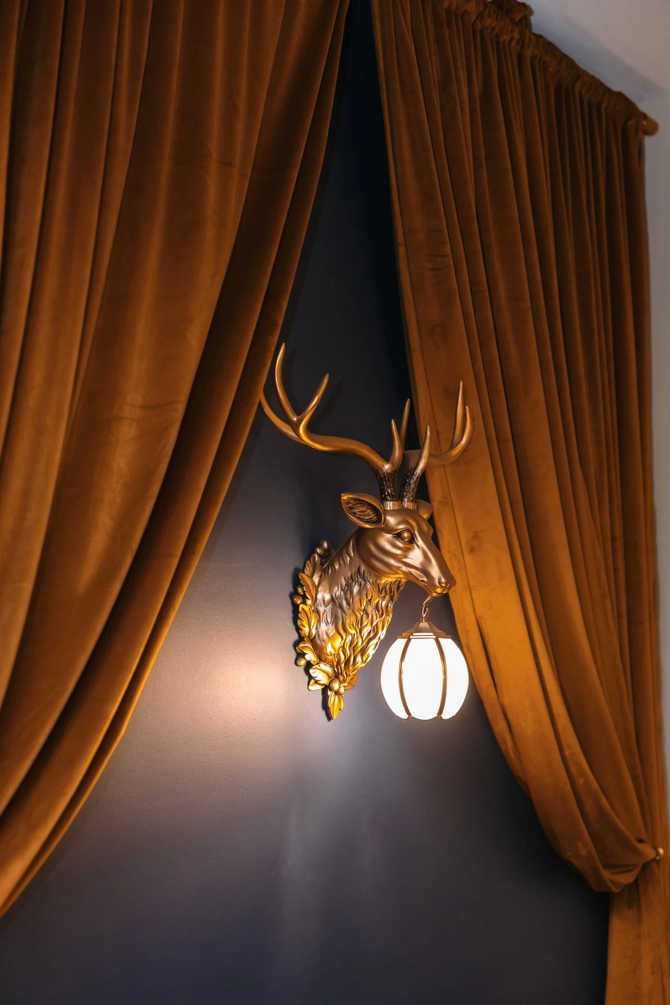 A gold-colored deer head wall sculpture with antlers is mounted on a wall between brown curtains, with a hanging lantern-style light fixture below in Ramble Creek groom's quarters.