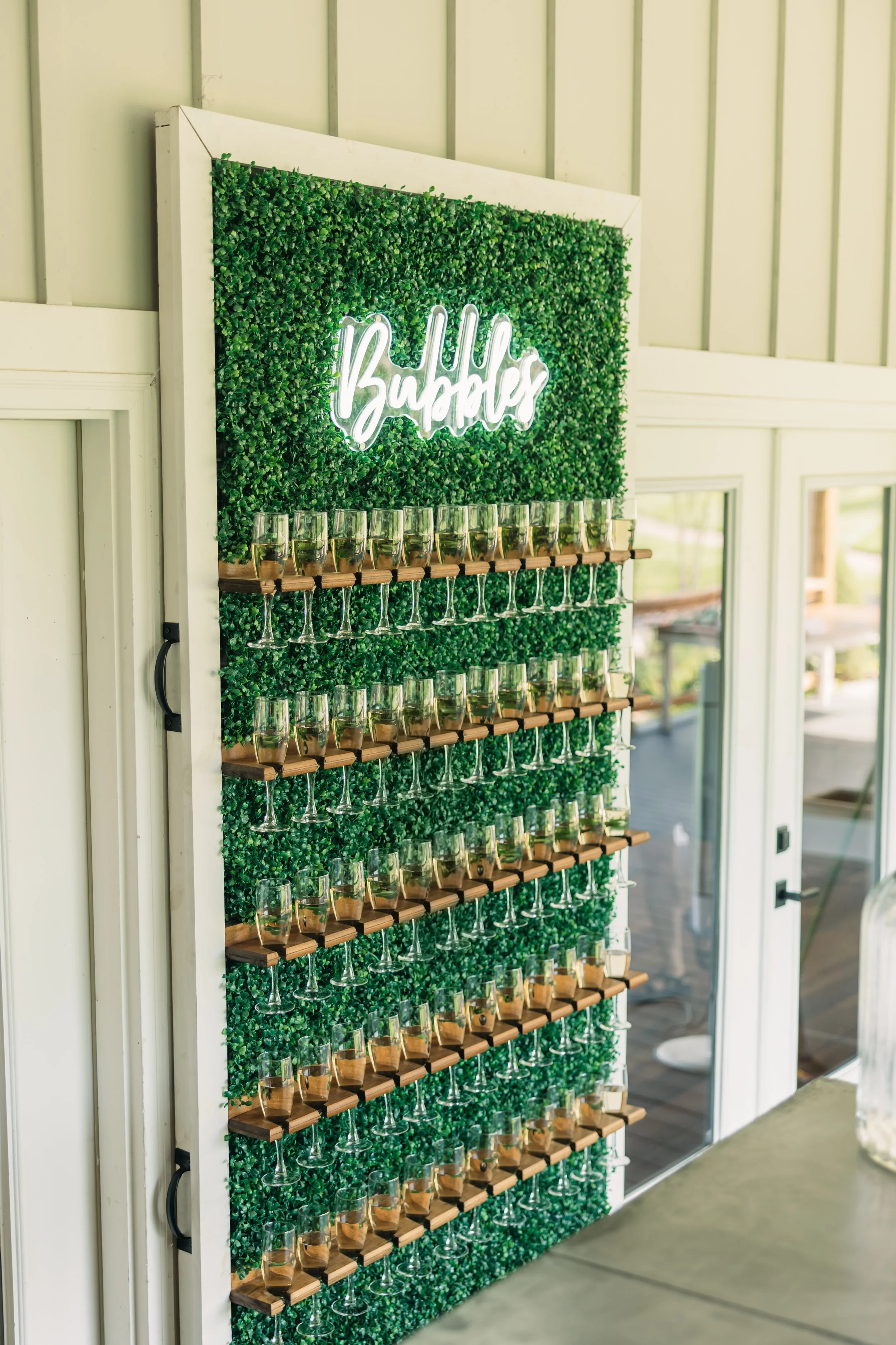 A decorative champagne wall with greenery and a neon sign that says 'Bubbles', holding rows of champagne glasses. Served during cocktail or dessert hour.