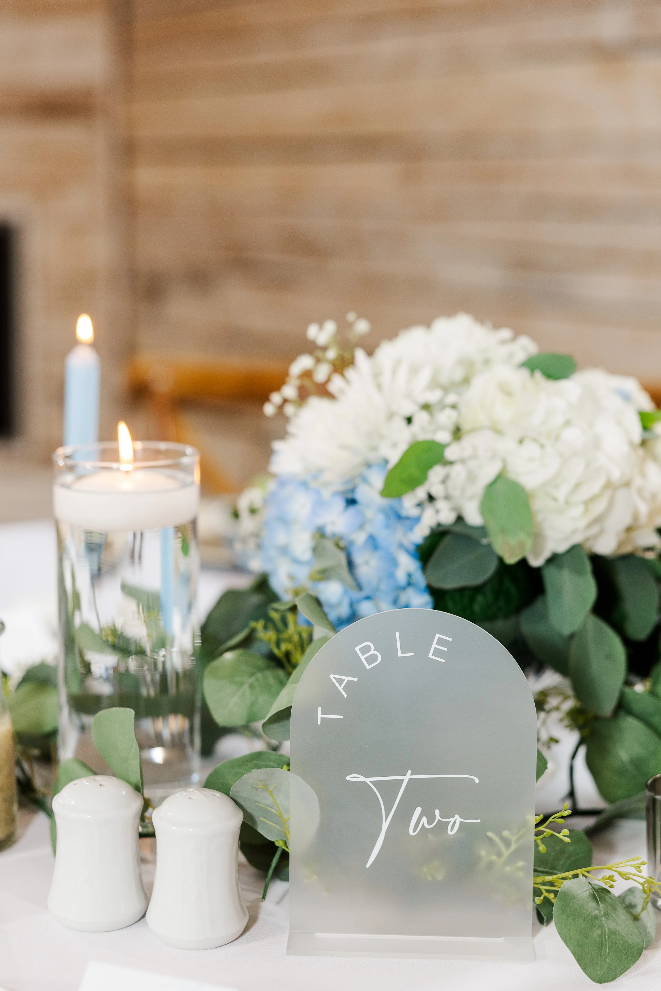 A wedding table centerpiece with white and light blue hydrangea flowers, greenery, three white salt and pepper shakers, three tall drinking glasses with lit candles inside, and a table number sign that reads 'Table Two'.