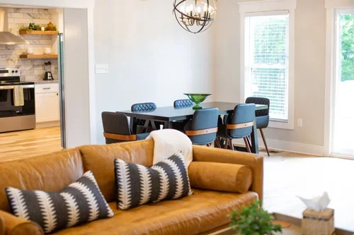 Dining area inside the Villa at Ramble Creek, featuring a dark dining table, upholstered chairs, chandelier, leather sofa, and open view into the kitchen.