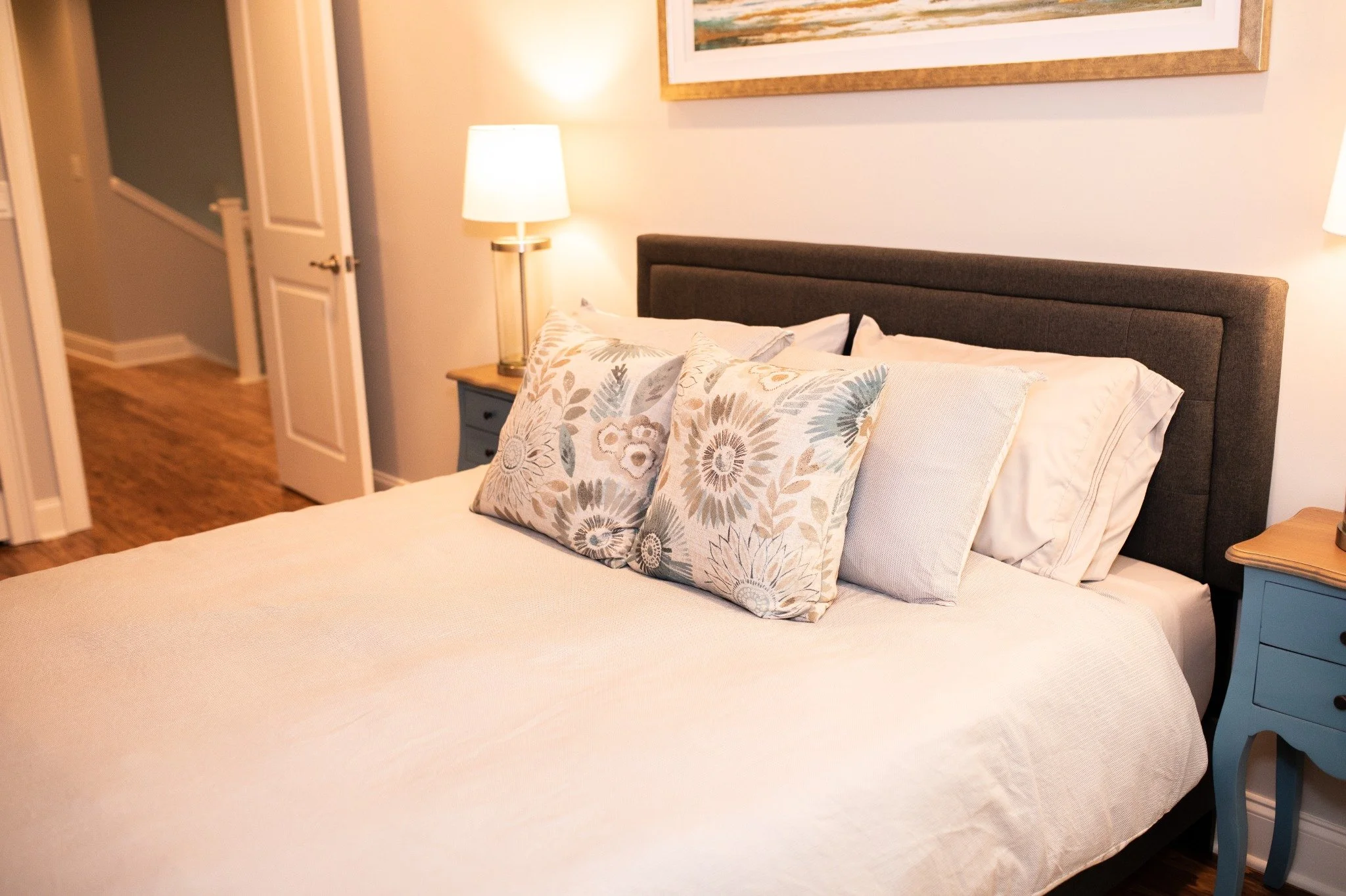 Downstairs bedroom at the Manor onsite accommodation at Ramble Creek, featuring a bed with white bedding, floral accent pillows, bedside lamps, blue nightstands, and warm wood floors.