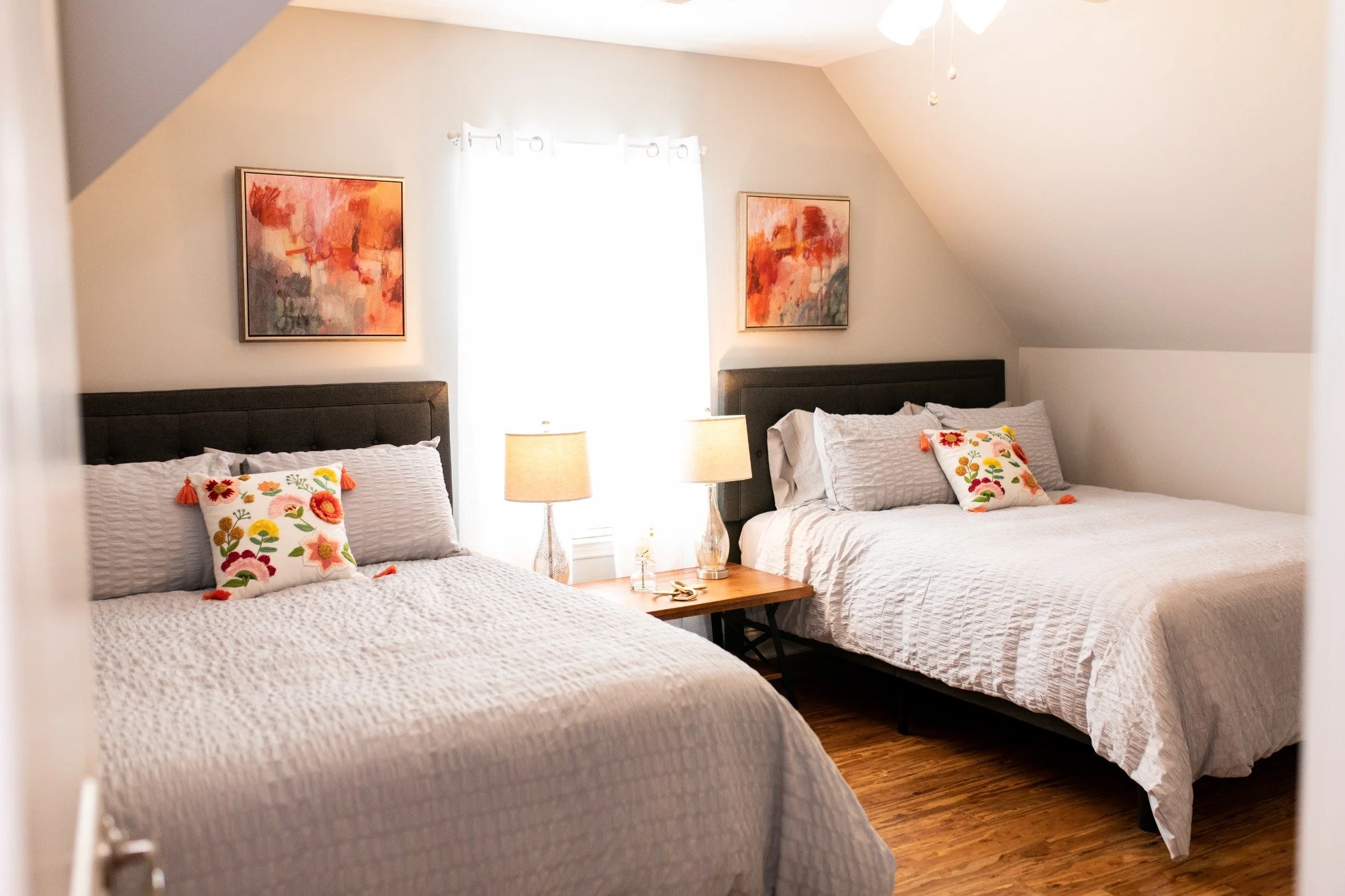 Upstairs bedroom at the Manor onsite accommodation at Ramble Creek, featuring two beds with white bedding, floral accent pillows, bedside lamps, warm wood floors, and soft neutral walls.