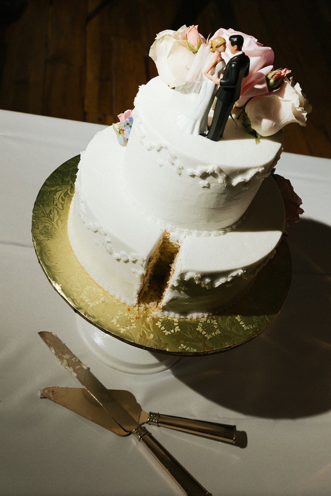 Wedding cake topped with flower decorations and bride and groom figurines, with a slice cut out, on a gold cake board, on a white table with cake serving utensils nearby.