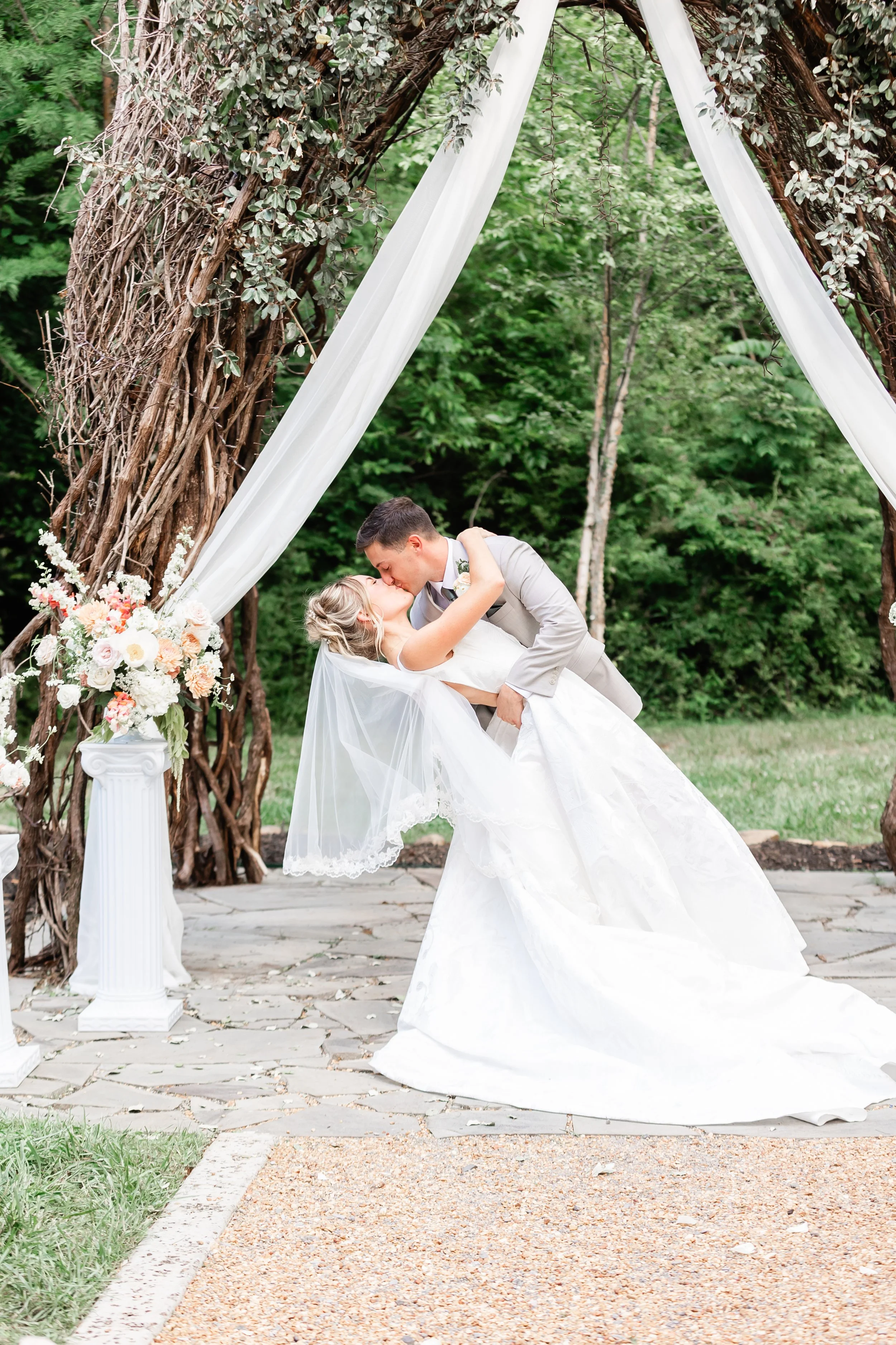 Bride and groom kissing beneath a romantic vine arbor draped with white fabric and pastel florals during an outdoor Tennessee wedding ceremony
