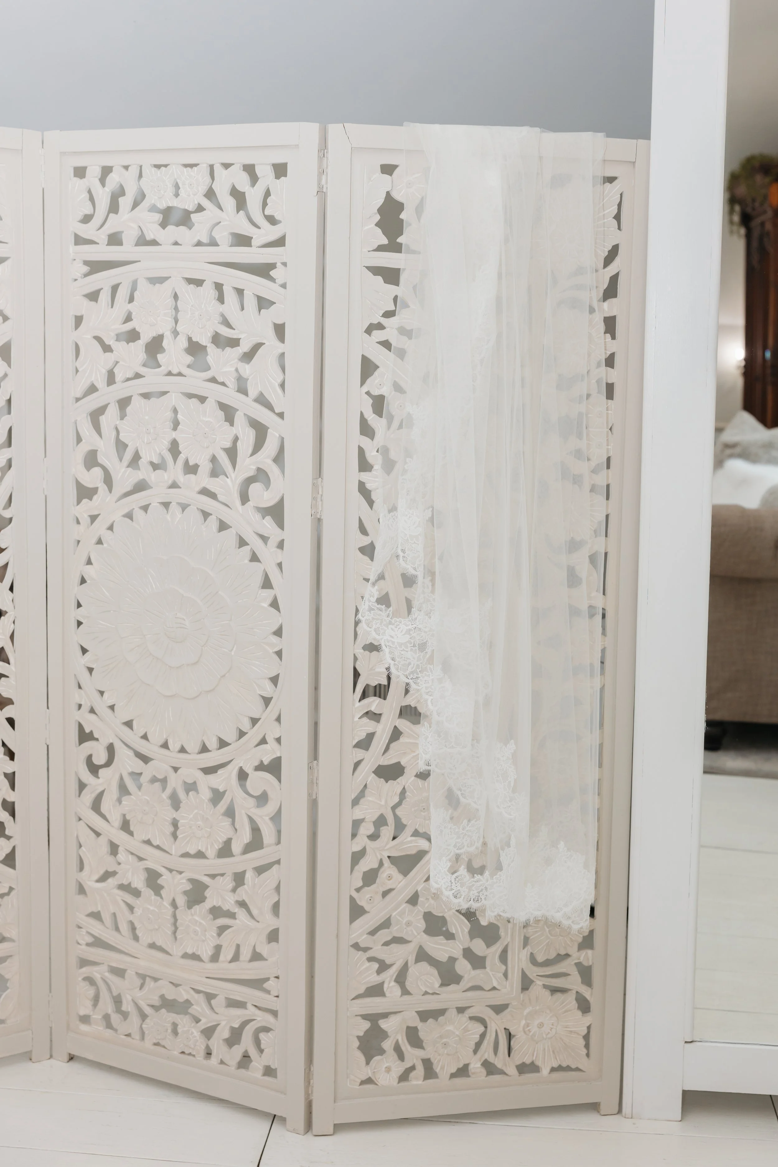 White decorative room divider with intricate floral and leaf cutout patterns, with a sheer white lace curtain hanging on one side, where bridesmaids hide their bags out of sight for wedding day pictures.