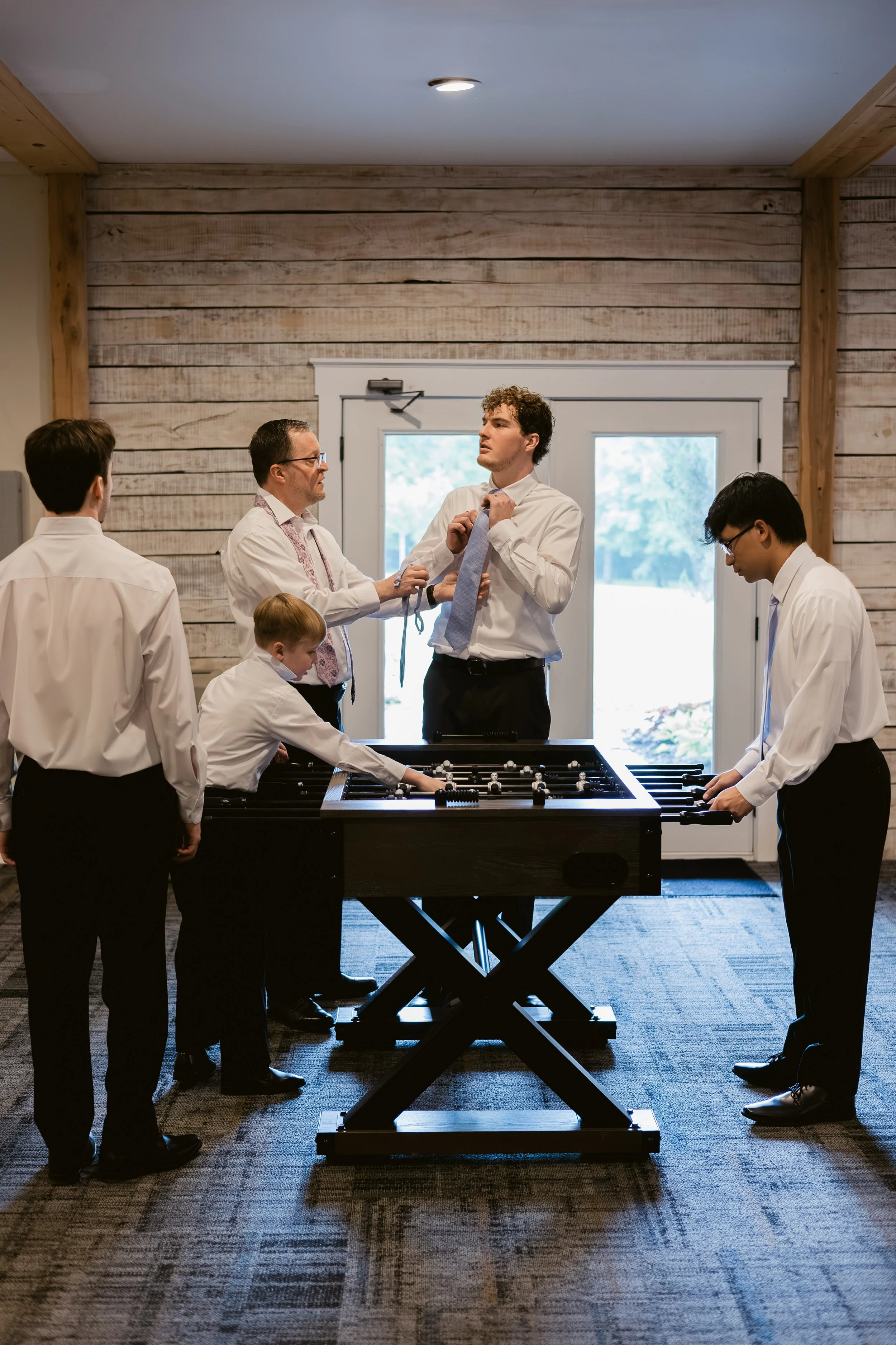 A groom and his groomsmen, all dressed in white shirts and black pants, playing foosball in a room with wooden walls and a large window. One man is adjusting his tie, while others are engaged in the game or watching.
