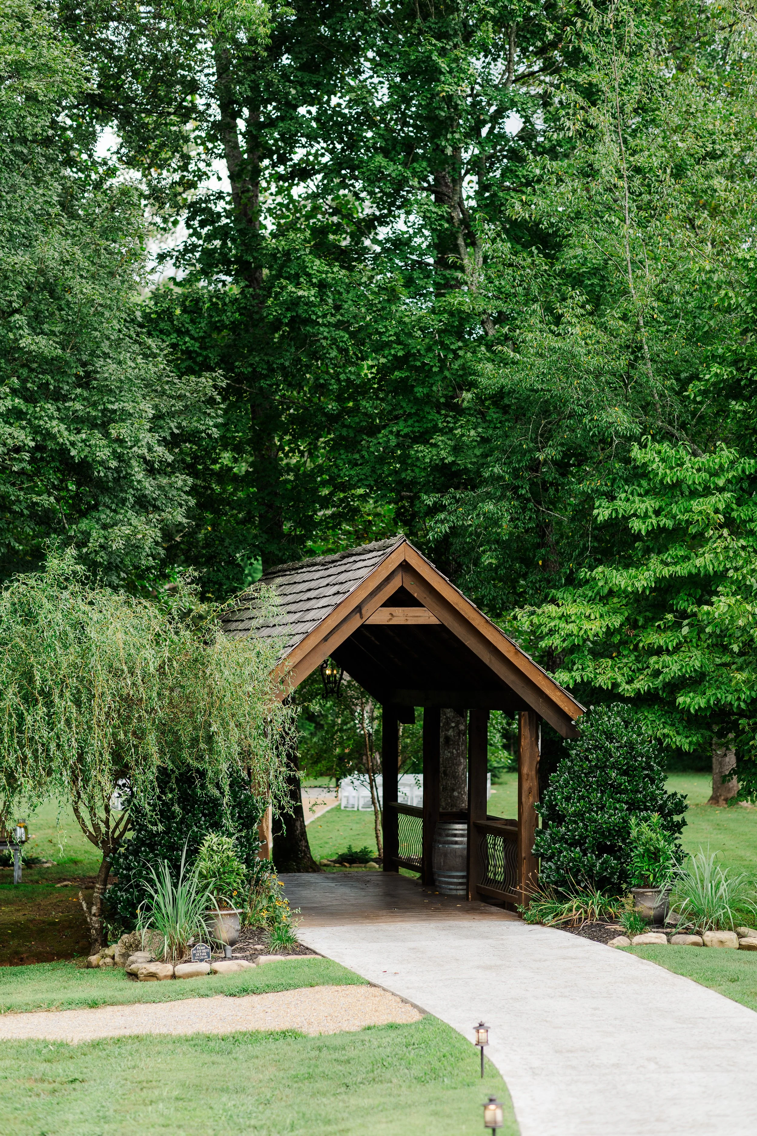 A wooden covered bridge with a shingled roof, situated in a lush green garden with various trees and shrubs, and a paved walkway leading up to it decorated with small lanterns.