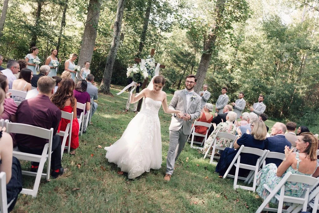 Bride and groom walking down the aisle at an outdoor wedding ceremony with guests seated on either side and bridesmaids and groomsmen standing in the background among trees at Ramble Creek all-inclusive wedding venue.