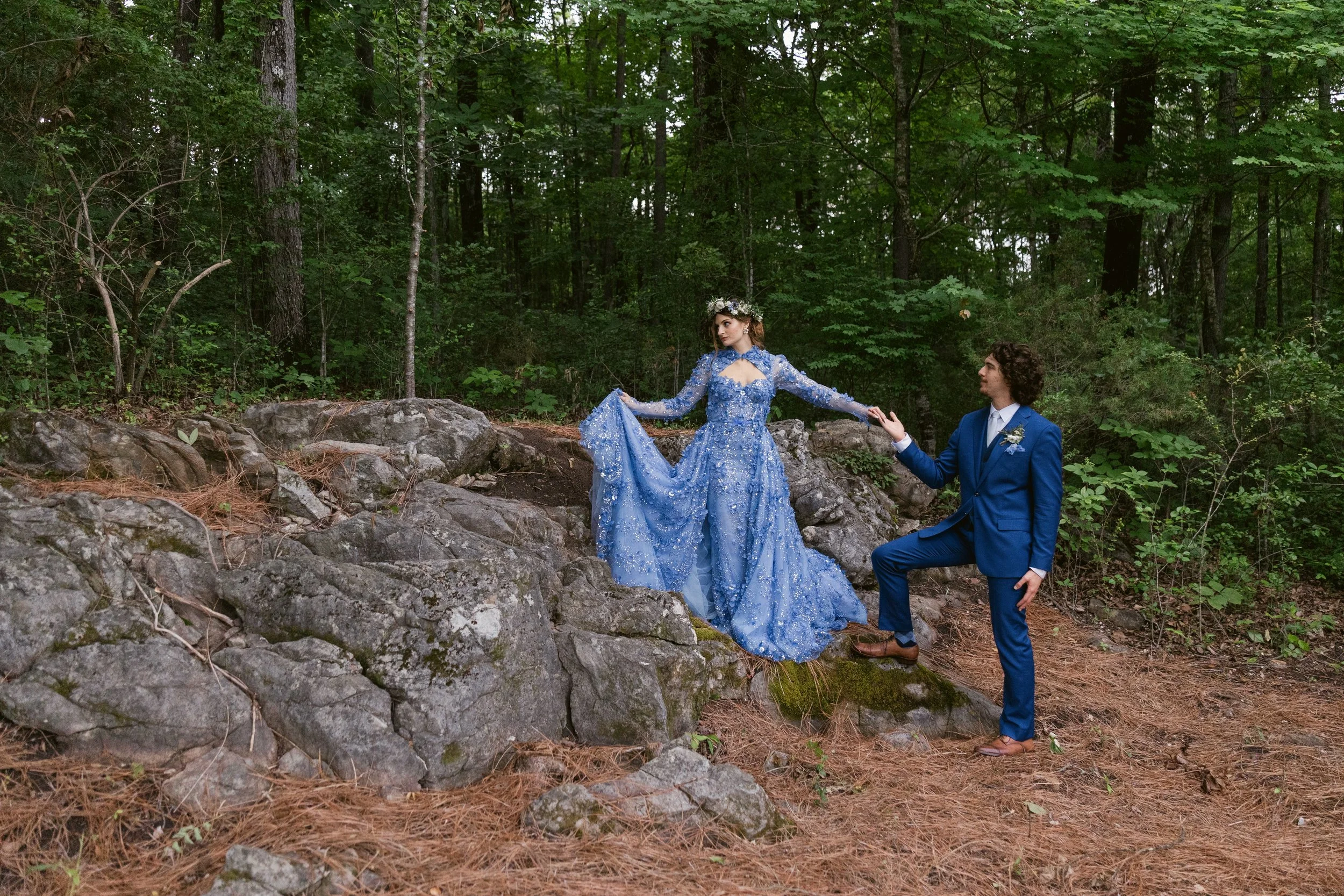 A bride in a blue, embellished gown and a floral crown sitting on rocks in a forest, holding hands with her groom in a blue suit, in a wedding or fantasy-themed photoshoot at Ramble Creek wedding venue near Chattanooga.