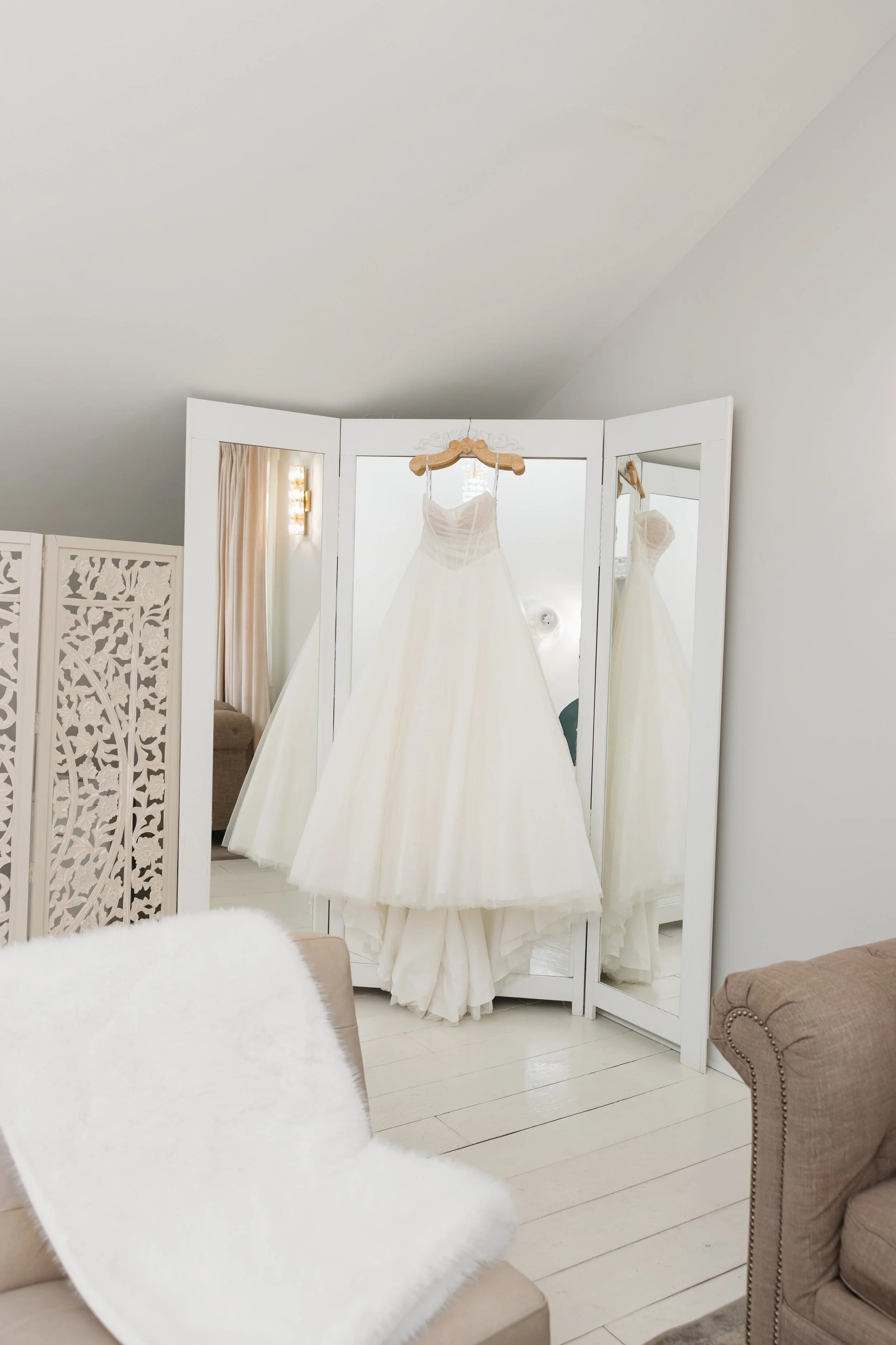 A white wedding dress hangs on a hanger in front of a full-length mirror in Ramble Creek bridal suite. Some furniture, including a taupe armchair with nailhead trim and a beige sofa, is visible in the room, perfect for a bride and her bridesmaids.