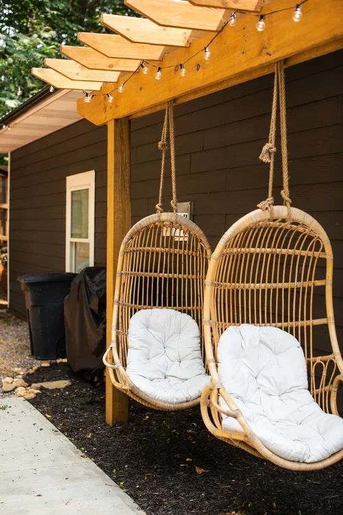 Two hanging rattan swings with white cushions under a wooden pergola with string lights, next to a house with dark siding and a window.