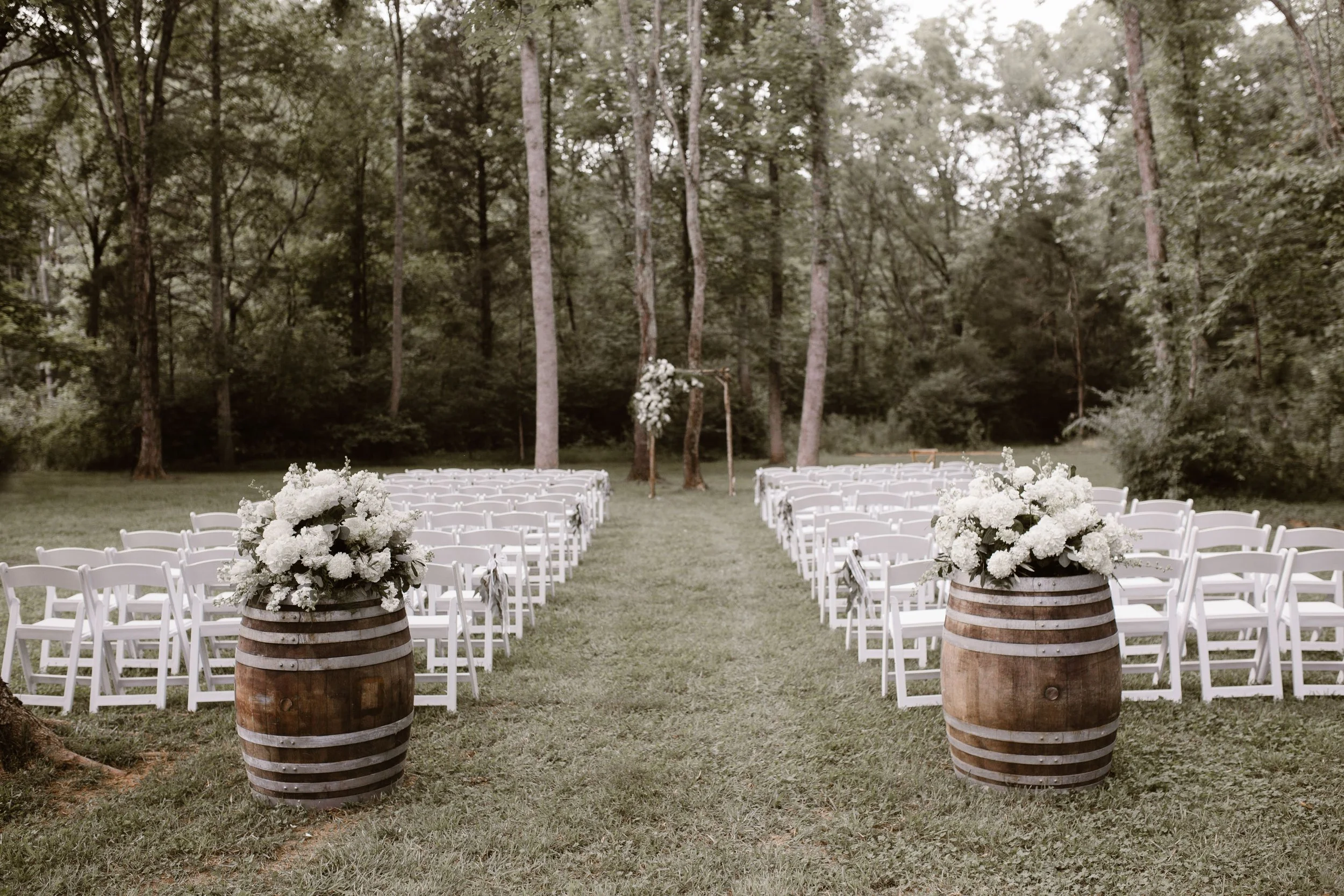 Outdoor wedding setup with white chairs arranged in rows, decorated with large white floral arrangements in wooden barrels, with a forest background at Ramble Creek East Tennessee wedding venue.