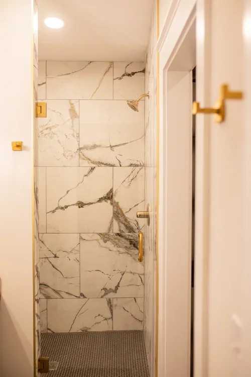 A small, tiled shower stall with white and gray marble-patterned tiles, gold fixtures, and a dark textured floor mat inside. One of three accommodations at Ramble Creek Tennessee wedding venue with on-site lodging.