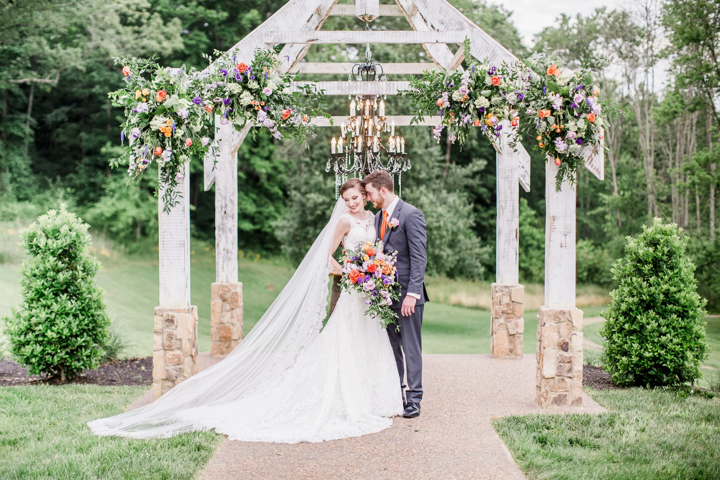 A bride and groom standing under a rustic wooden wedding arch decorated with colorful flowers and greenery, with a chandelier hanging above them, outdoors in a garden setting at Ramble Creek wedding venue in Athens, TN.