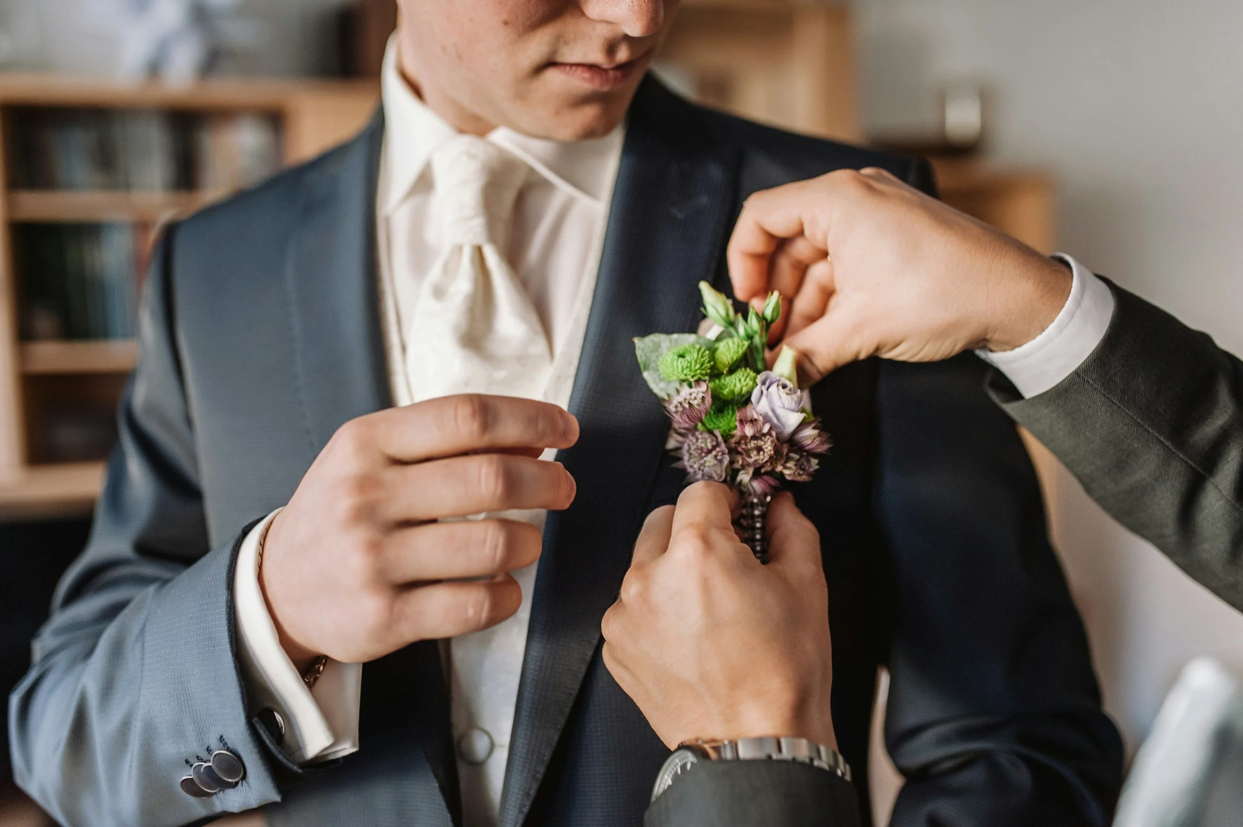 Close-up of a groom’s boutonniere featuring lavender blooms, green textured florals, and soft greenery being pinned onto a navy suit jacket at Ramble Creek.