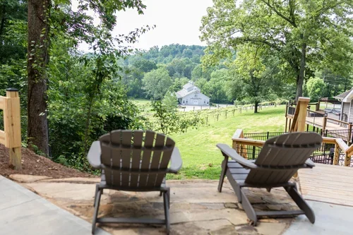 Two Adirondack chairs on a wooden deck overlooking a lush green wedding estate with trees, a white indoor reception/ceremony space, and a vineyard. One of three accommodations at Ramble Creek Tennessee wedding venue with on-site lodging.