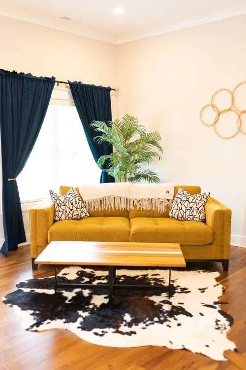 Living room with a yellow sofa, black and white patterned pillows, a throw blanket, a wooden coffee table, a cowhide rug, dark blue curtains, a green potted plant, and wall decor of interconnected gold rings.