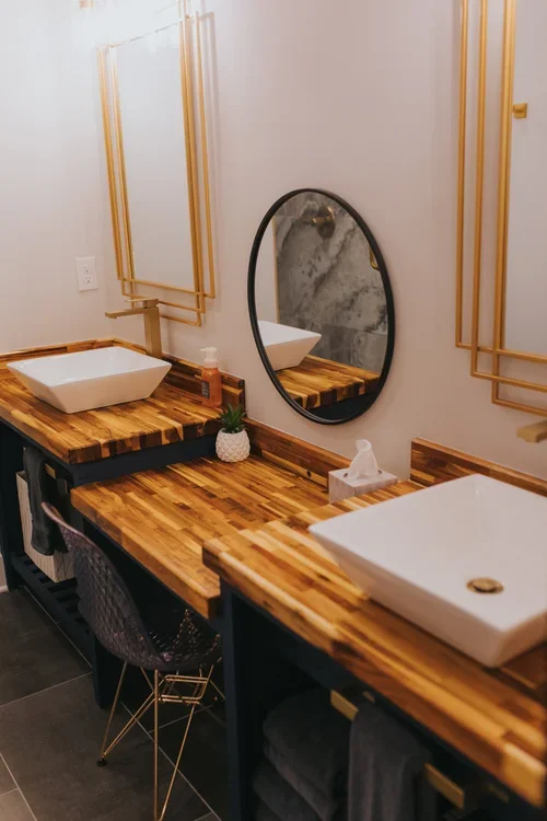 Modern bathroom vanity with two vessel sinks, round and rectangular, on a wooden countertop. There is a mirror above each sink and decorative golden wall frames. A small potted plant and tissue box are on the counter.
