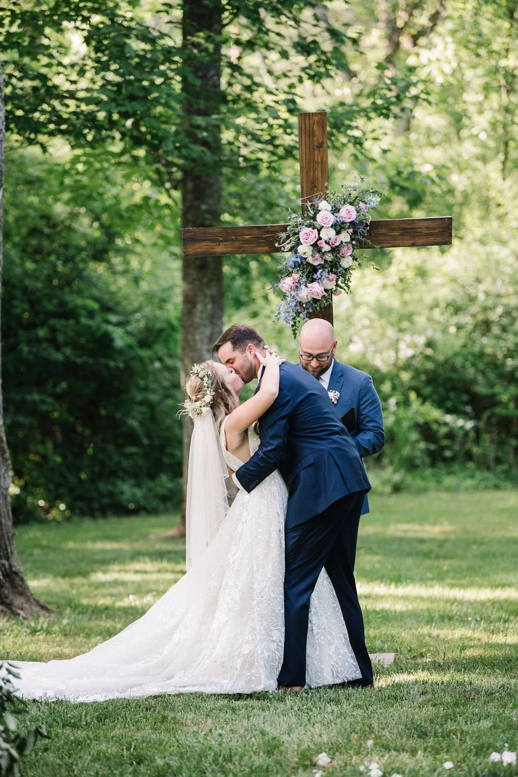 A wedding ceremony outdoors with a bride and groom kissing in front of a wooden cross decorated with pink and purple flowers, accompanied by an officiant, in a lush green wooded setting at Ramble Creek all-inclusive wedding venue.