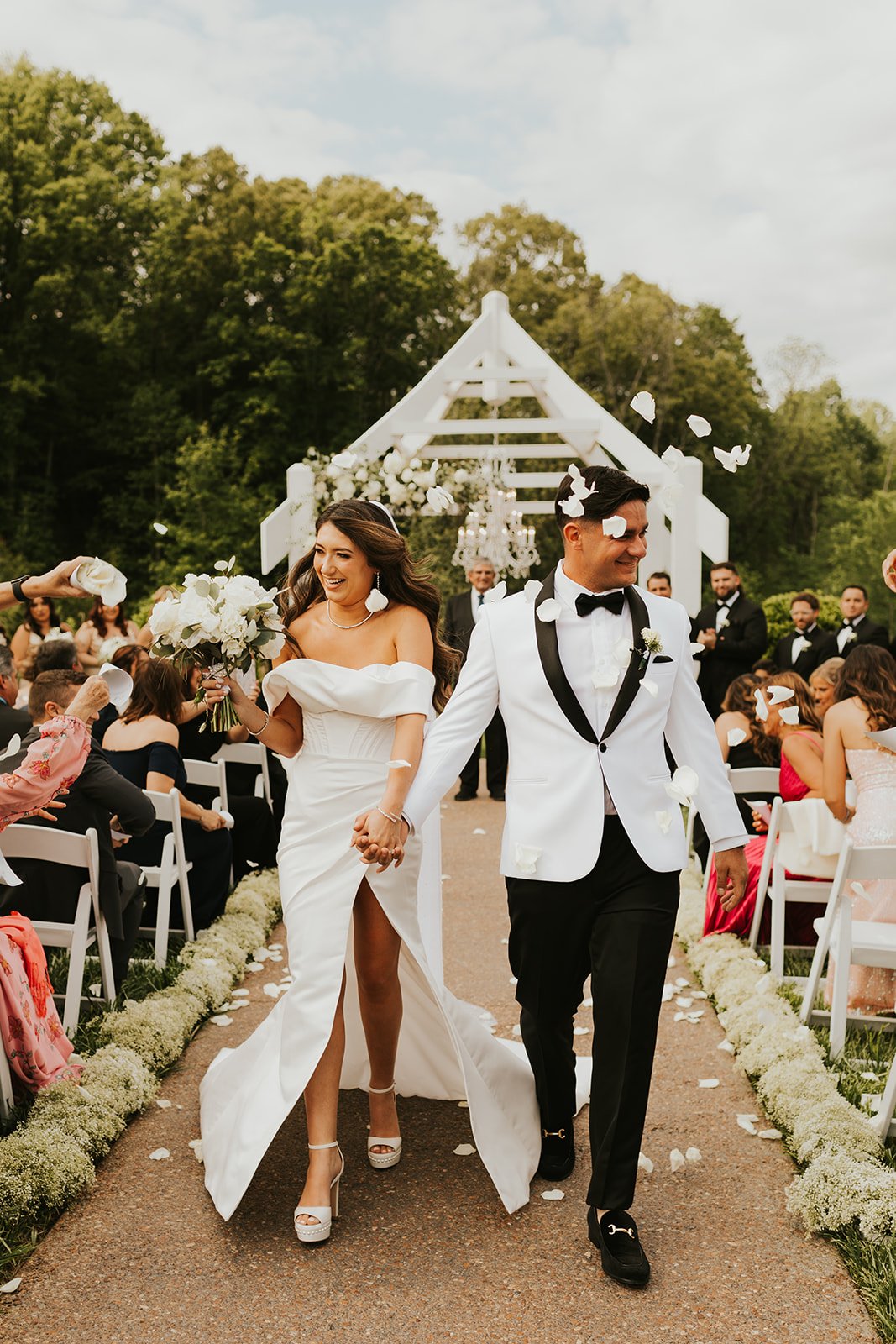 Bride and groom holding hands walking down aisle at Ramble Creek East Tennessee outdoor wedding, guests cheering, white petals and flowers on path, decorated white arbor in background.
