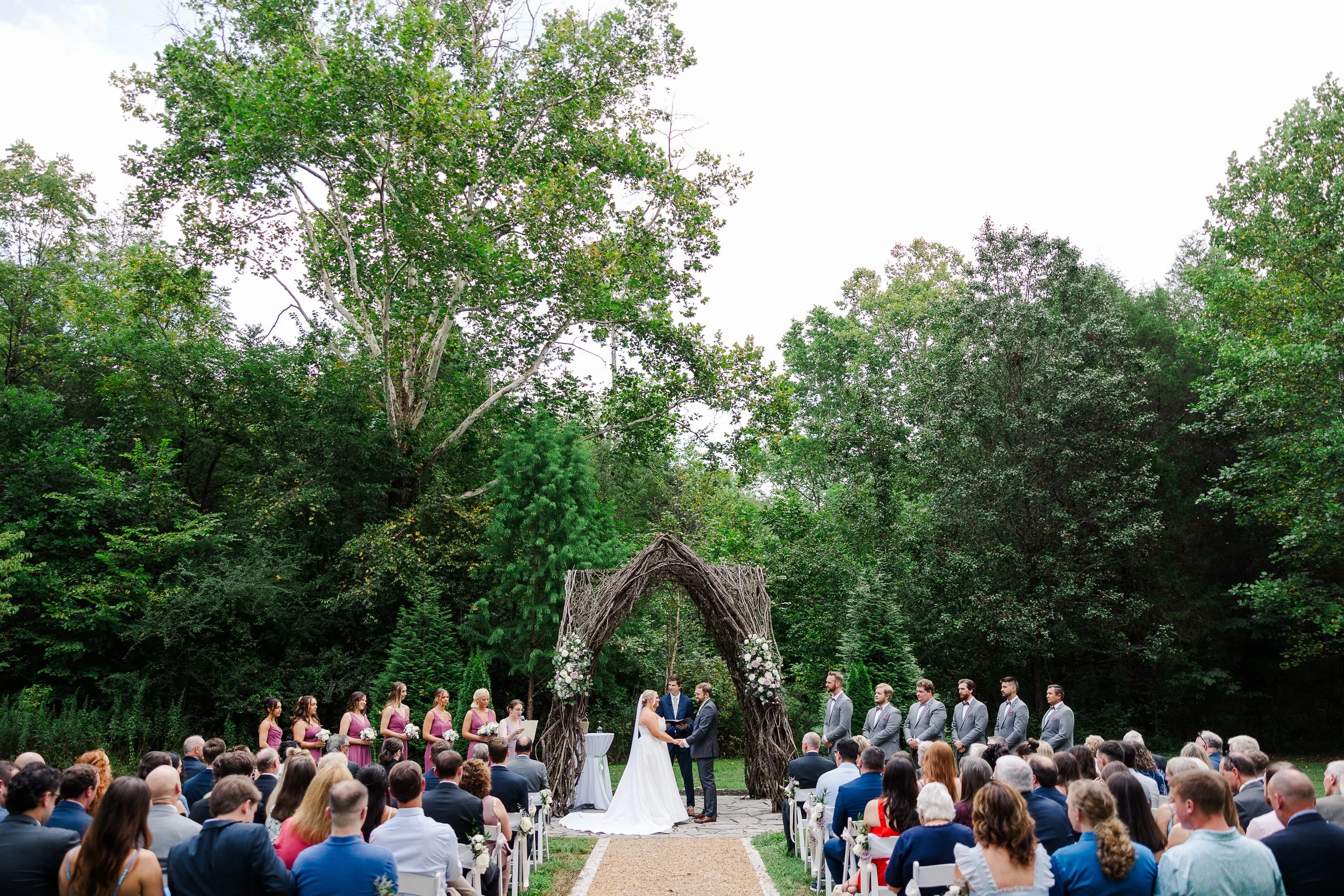Outdoor summer wedding ceremony at Ramble Creek with guests gathered beneath lush Tennessee trees and a natural vine arbor.