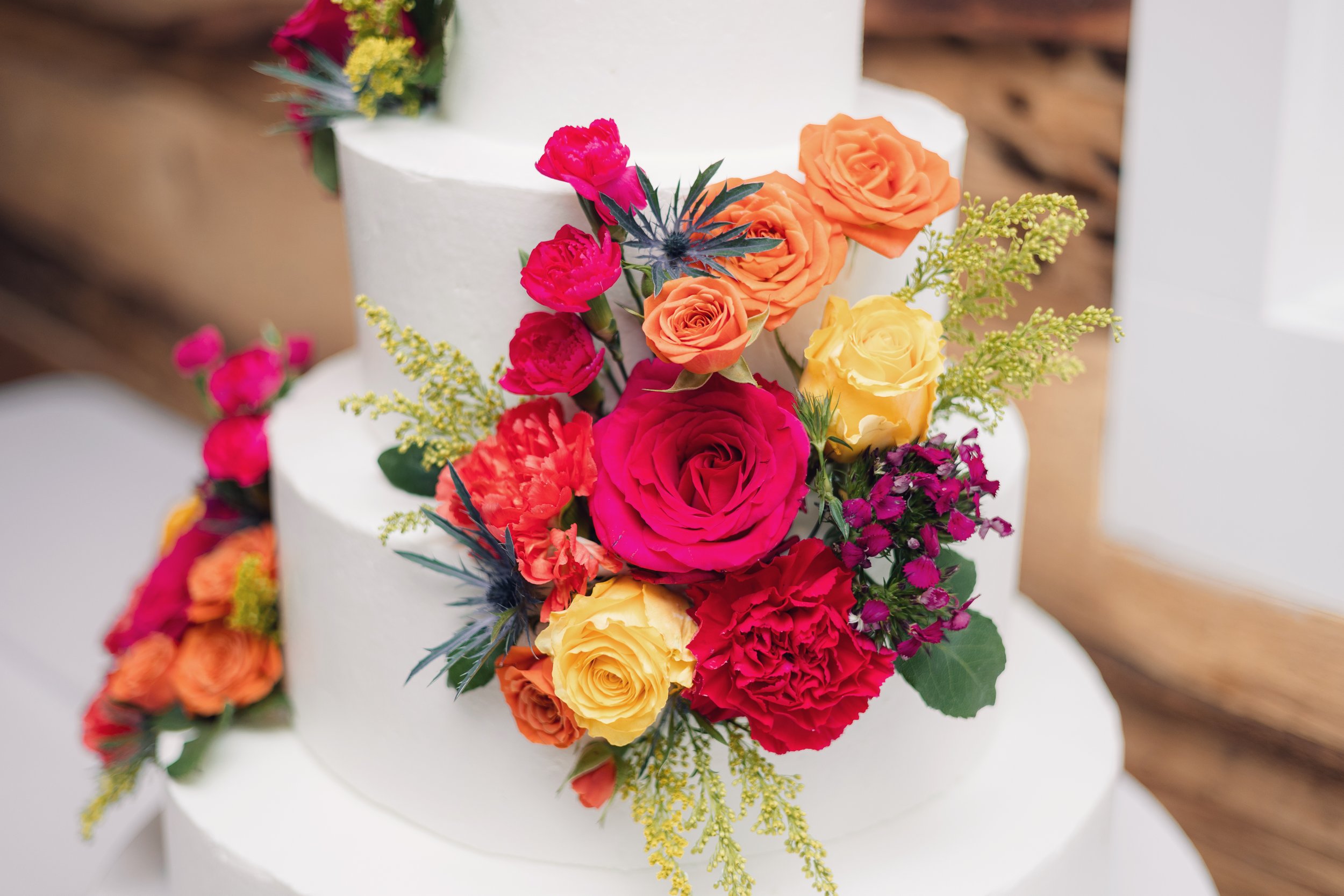 Close-up of a tiered white wedding cake decorated with a colorful bouquet of flowers including roses, carnations, and greenery, showcasing Ramble Creek's in-house floral and bakery services.