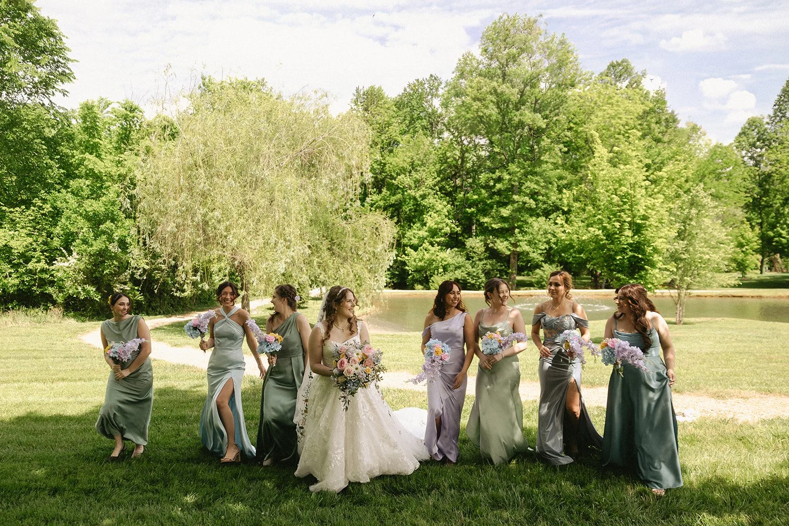 Bride with bridesmaids in soft green and lavender dresses by the pond at Ramble Creek during a colorful May spring wedding in Tennessee.