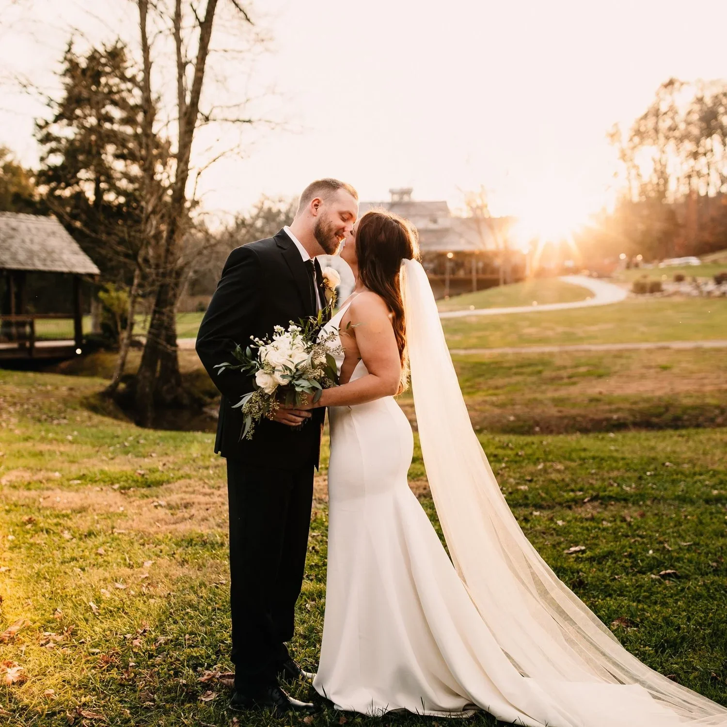 A bride and groom stand close together in a park during sunset, with trees and a building in the background. The bride is wearing a white wedding dress and veil, holding a bouquet of flowers, and the groom is in a black suit. They are smiling and touching foreheads.