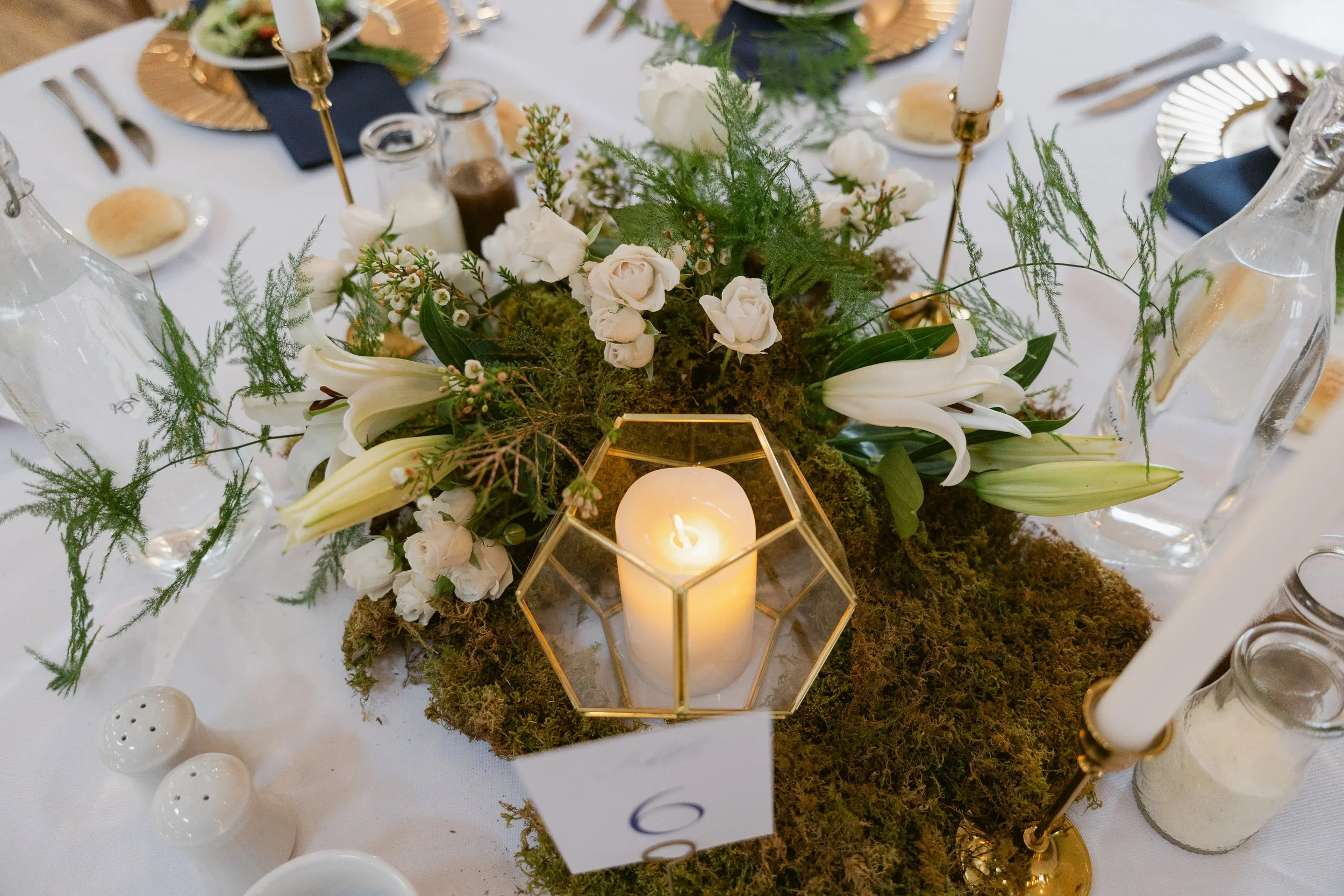 Candlelit floral reception centerpiece with white roses, greenery, and moss on a wedding table at Ramble Creek Wedding Venue