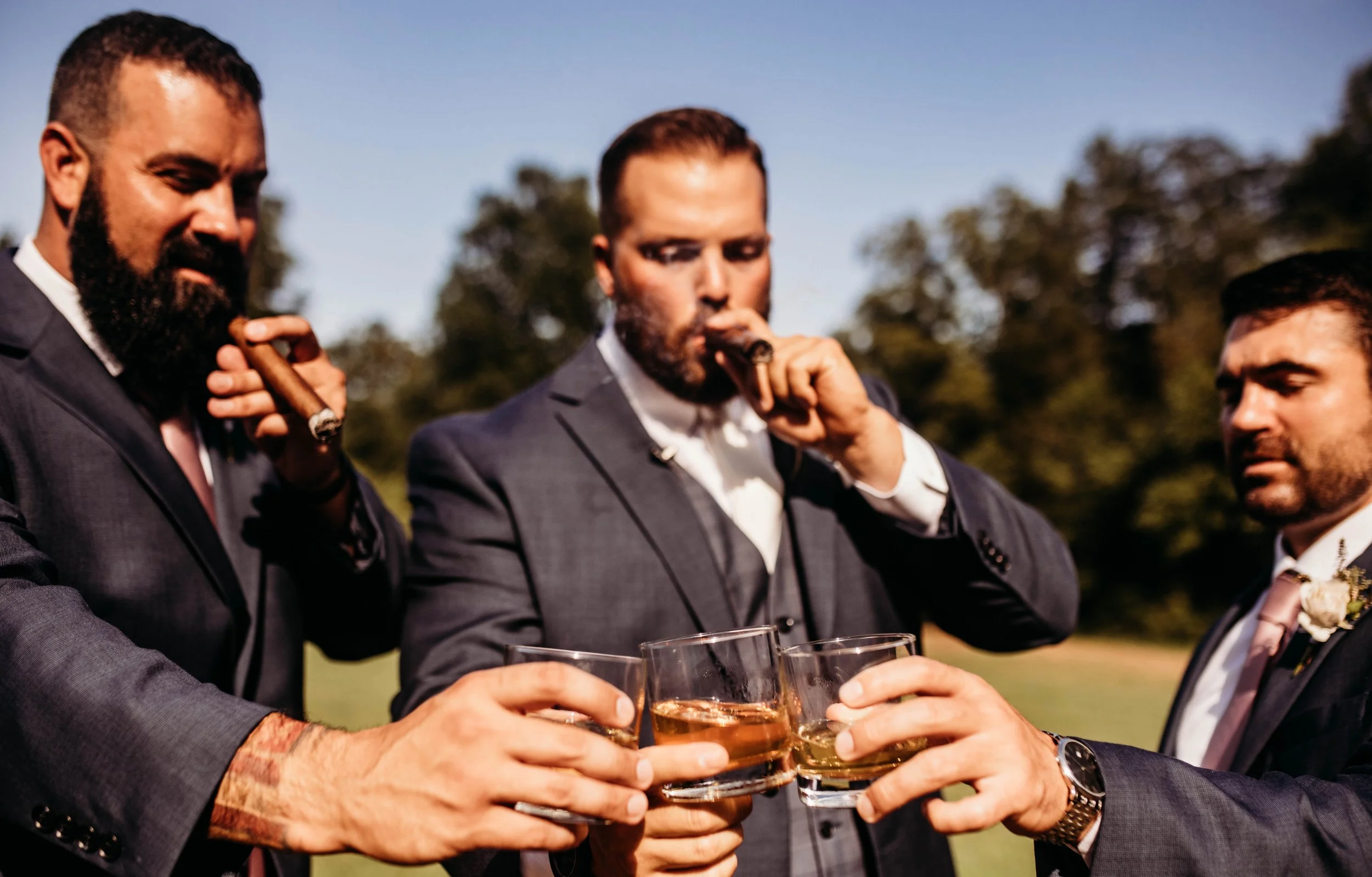 A group of men dressed in suits, each holding a glass of whiskey, toasting outdoors on a sunny day before their friend is married at Ramble Creek destination wedding venue in East Tennessee.