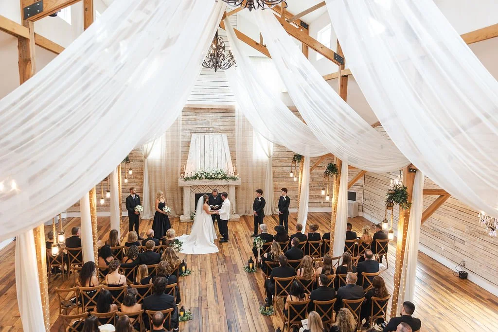 Indoor wedding ceremony in a rustic barn with wooden beams, white draping, and string lights, featuring the bride and groom exchanging vows in front of an officiant, surrounded by seated guests.