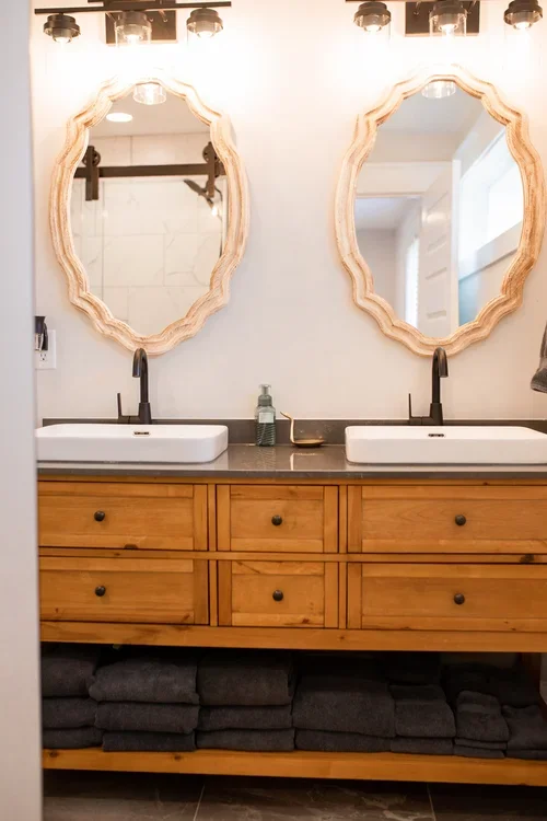 Primary bathroom inside the Villa at Ramble Creek, featuring a double vanity with vessel sinks, two decorative mirrors, wood cabinetry, black faucets, and gray towels.