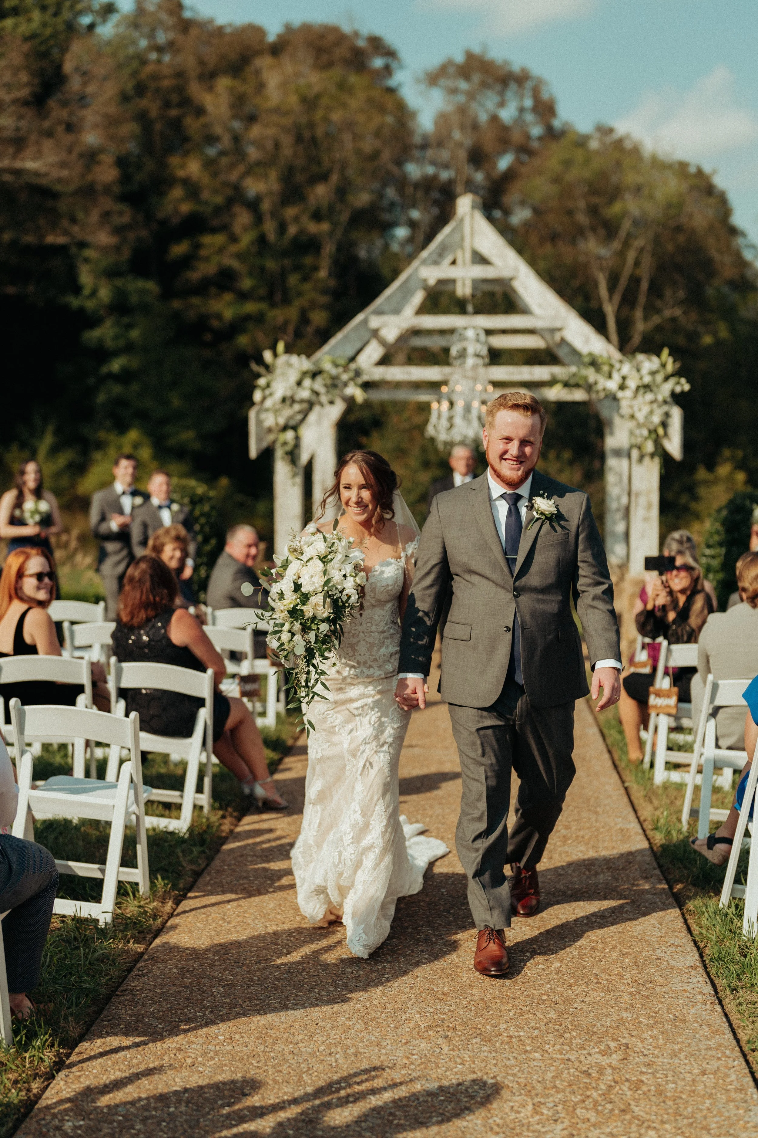 A newlywed couple walking down the aisle after their wedding ceremony outdoors. The bride is holding a large bouquet of white flowers and dressed in a lace wedding gown. The groom is wearing a gray suit with a dark tie. Guests are seated on white cha