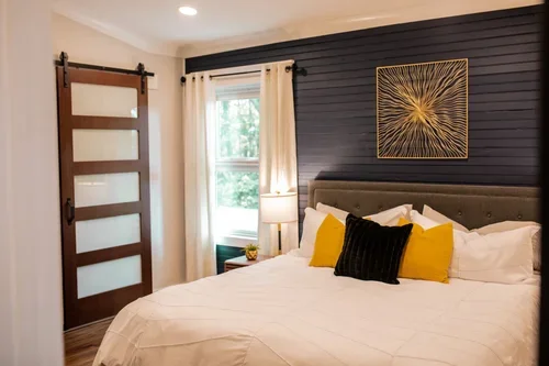 A bedroom with a large bed decorated with yellow and black pillows, a dark accent wall with a gold geometric artwork, a window with white curtains, a sliding barn door, and a nightstand with a lamp.