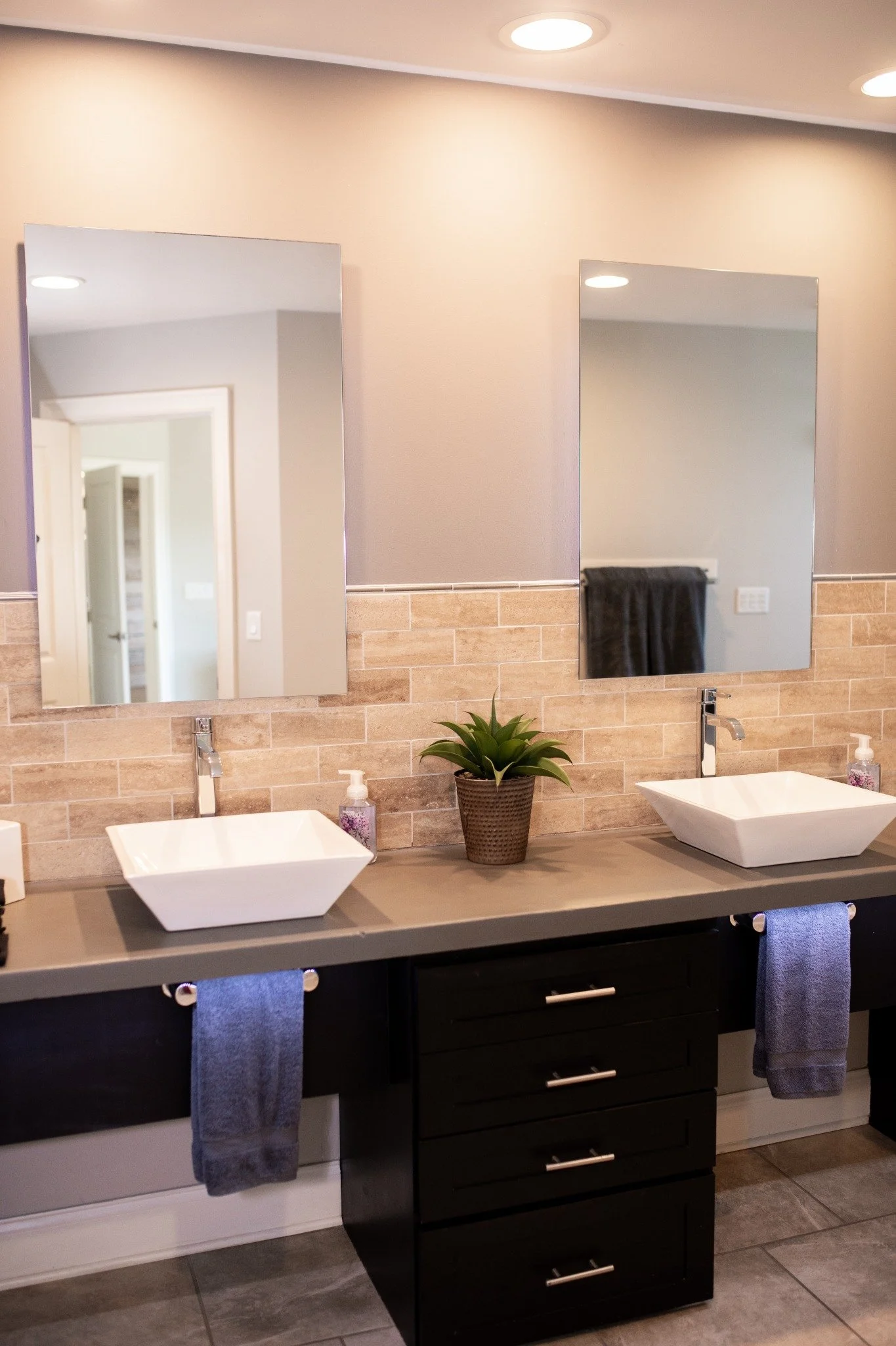 Double bathroom vanity with two vessel sinks, a potted plant between the mirrors, and towels hanging underneath.
