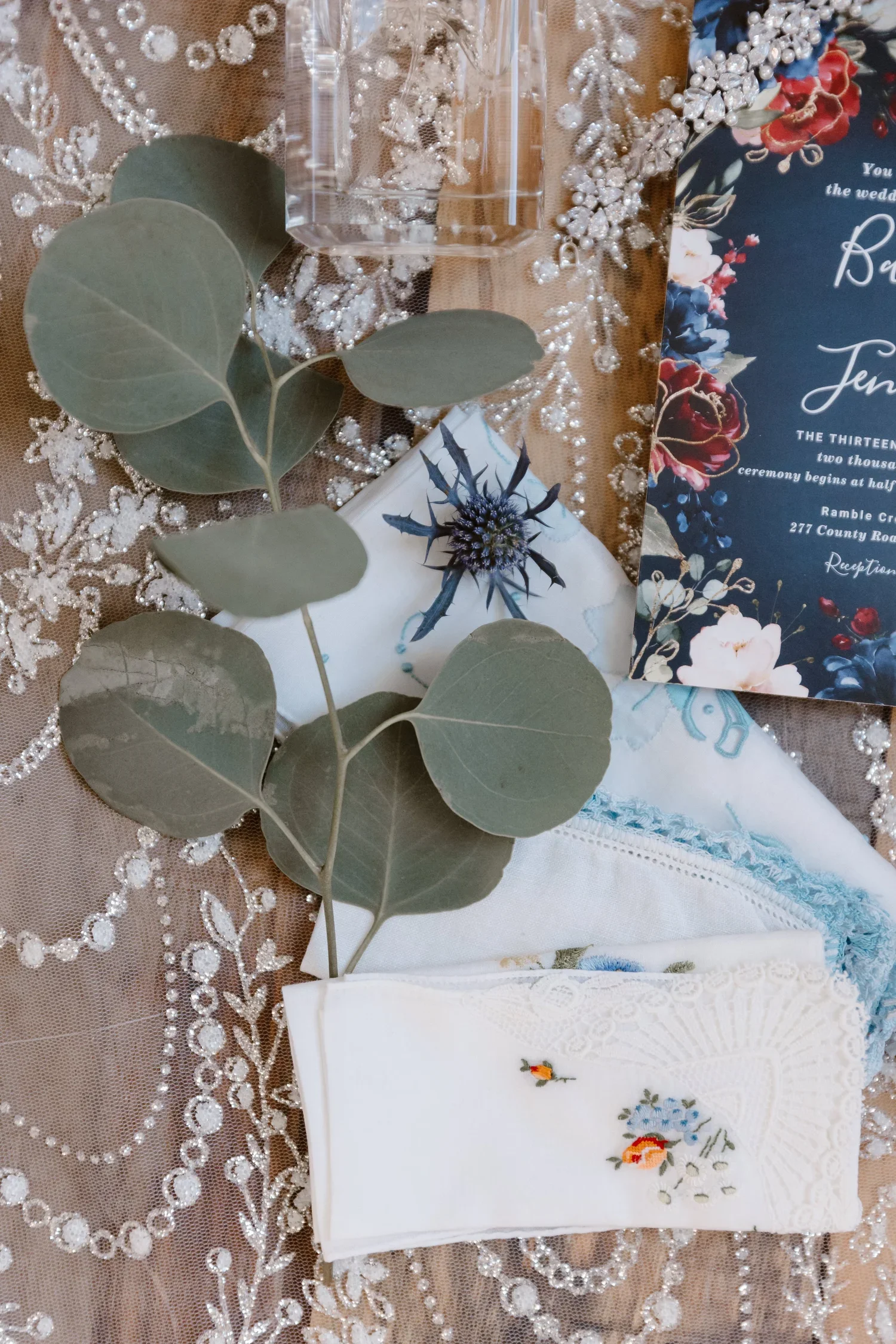Wedding detail flat lay with eucalyptus, blue thistle, embroidered handkerchiefs, lace, pearl accents, and a navy floral invitation for a Ramble Creek wedding