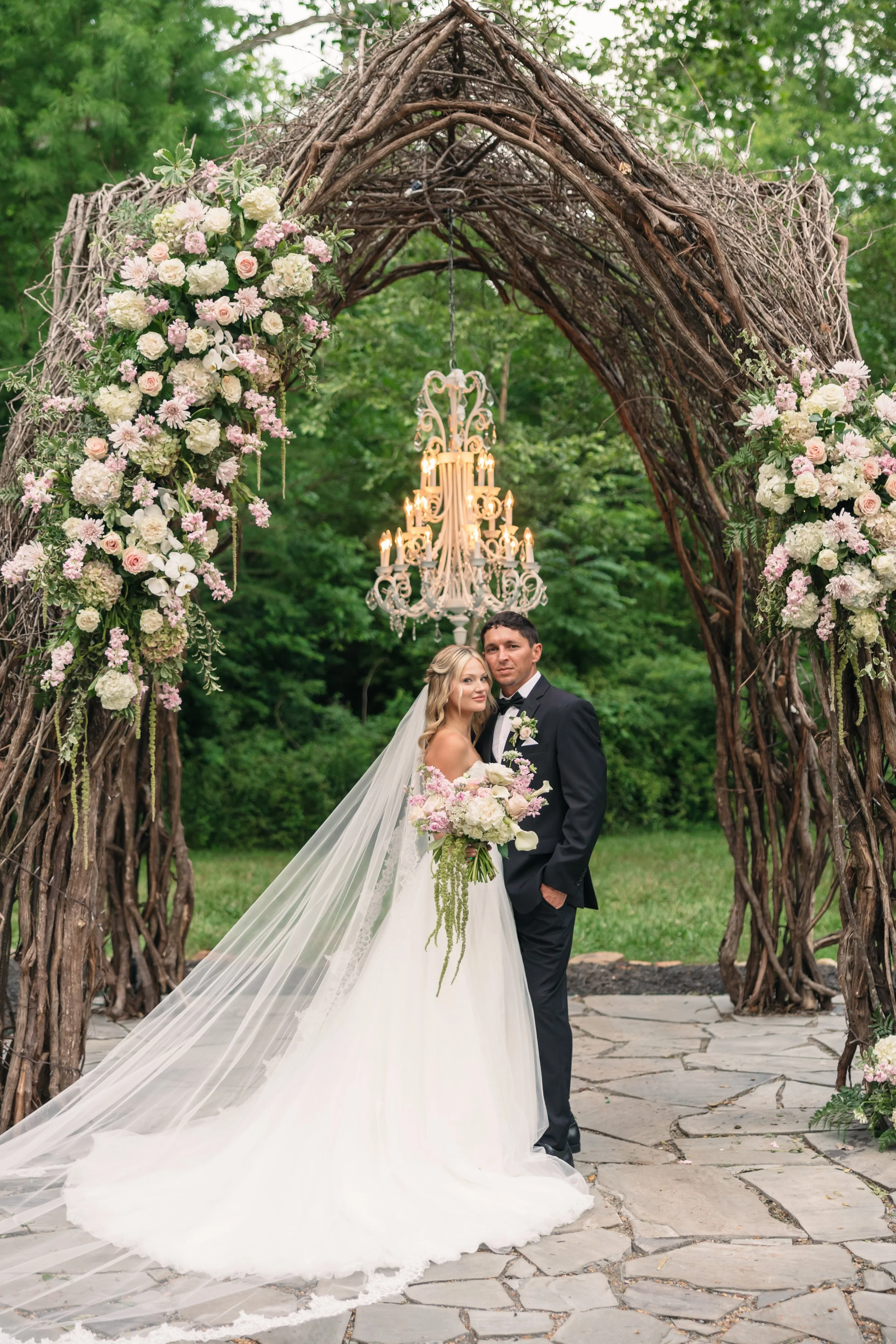 Bride and groom portrait beneath the woodland ceremony arch at Ramble Creek Wedding Venue, featuring cascading pastel florals, a chandelier, and stone walkway.