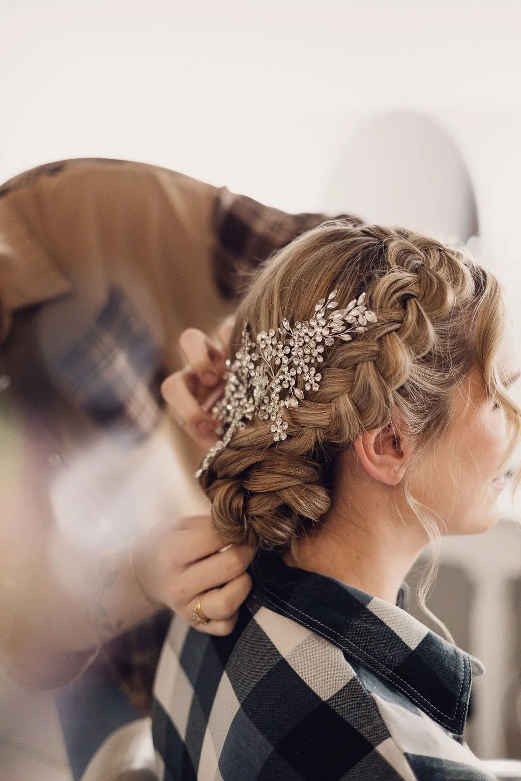 Bride’s braided low updo with a sparkling crystal hair vine, shown as the stylist secures final details for a polished wedding day look