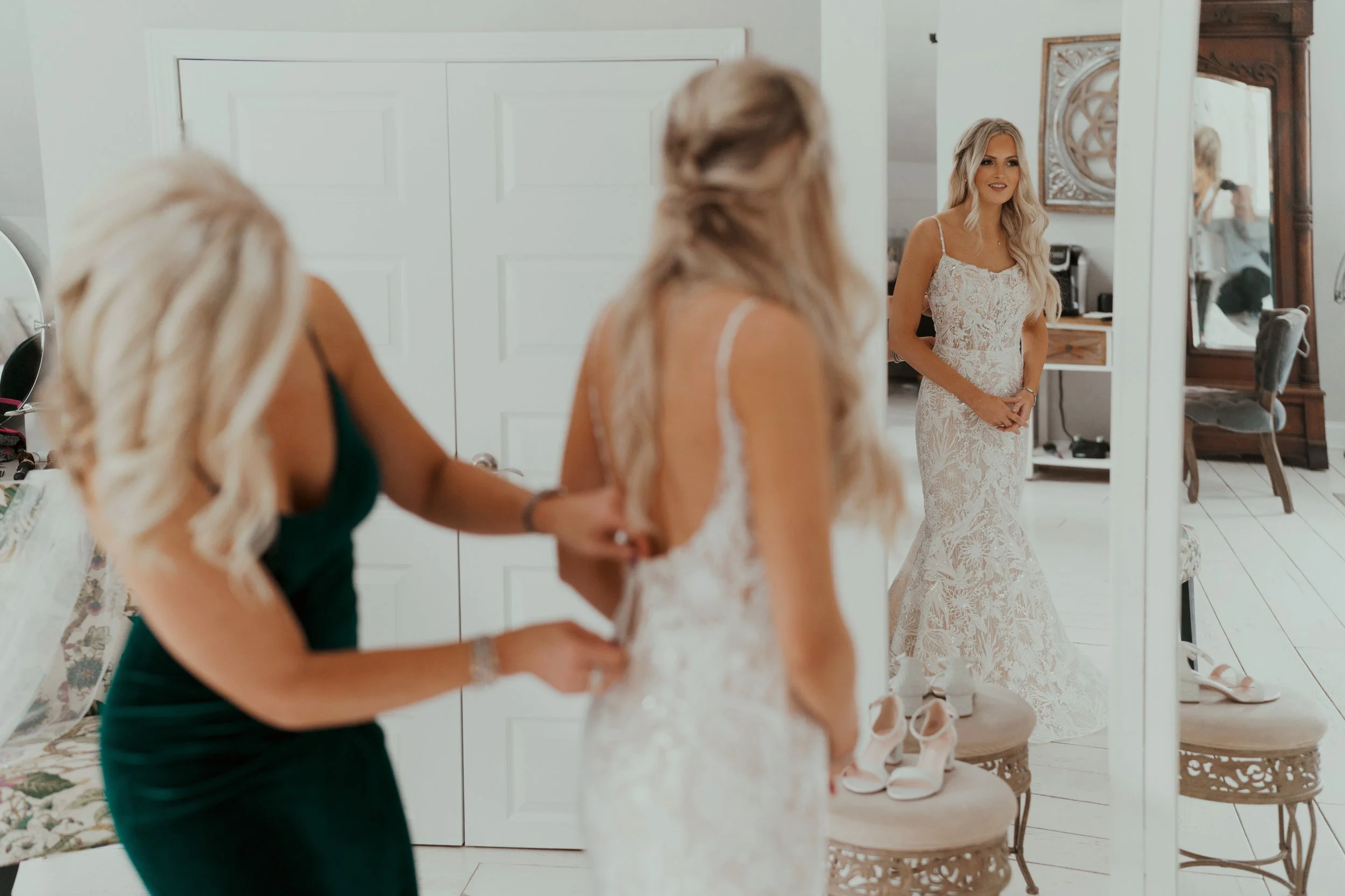 Bride getting ready in the Ramble Creek bridal suite as a bridesmaid buttons her lace wedding dress, with her reflection shown in the mirror
