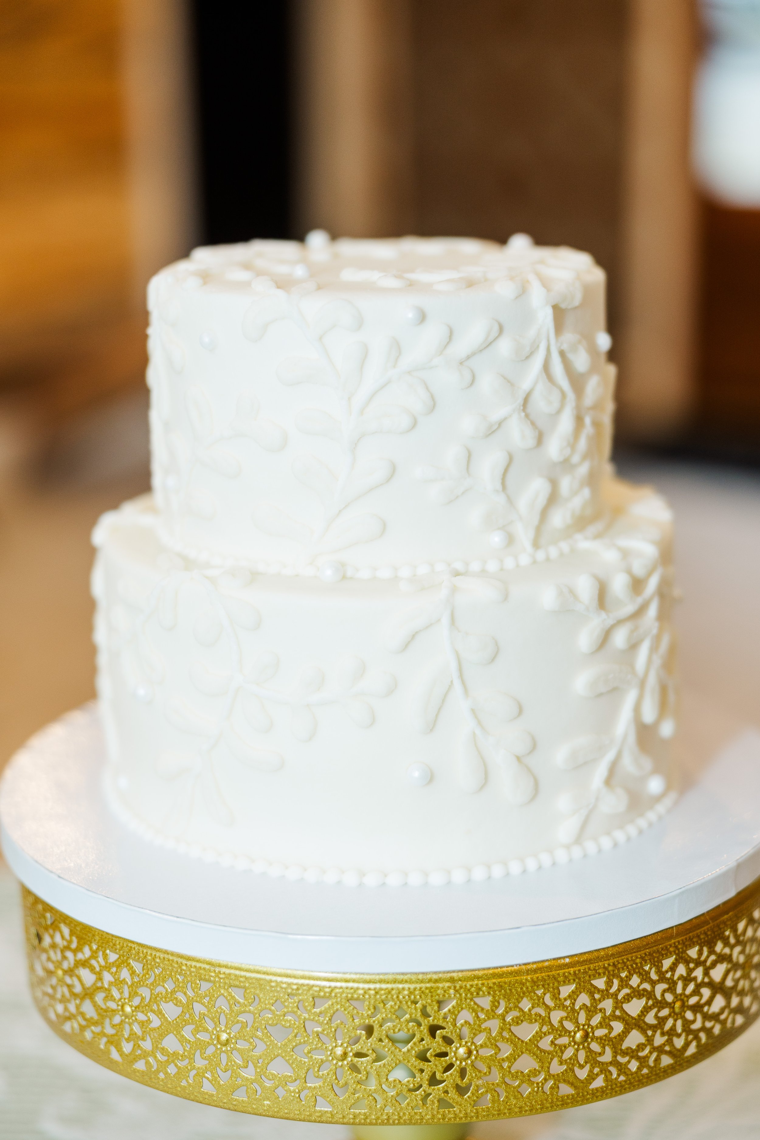 A two-tiered wedding cake decorated with white icing and intricate white piping patterns, placed on a white cake board with a gold decorative border, showcasing Ramble Creek in-house wedding bakery services.