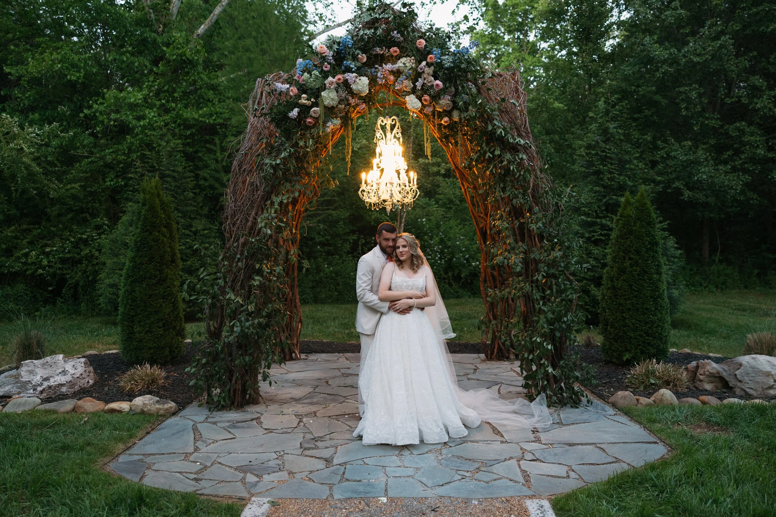 Bride and groom posing beneath Ramble Creek’s illuminated vine arbor with spring florals and a chandelier during an evening outdoor wedding in Tennessee.